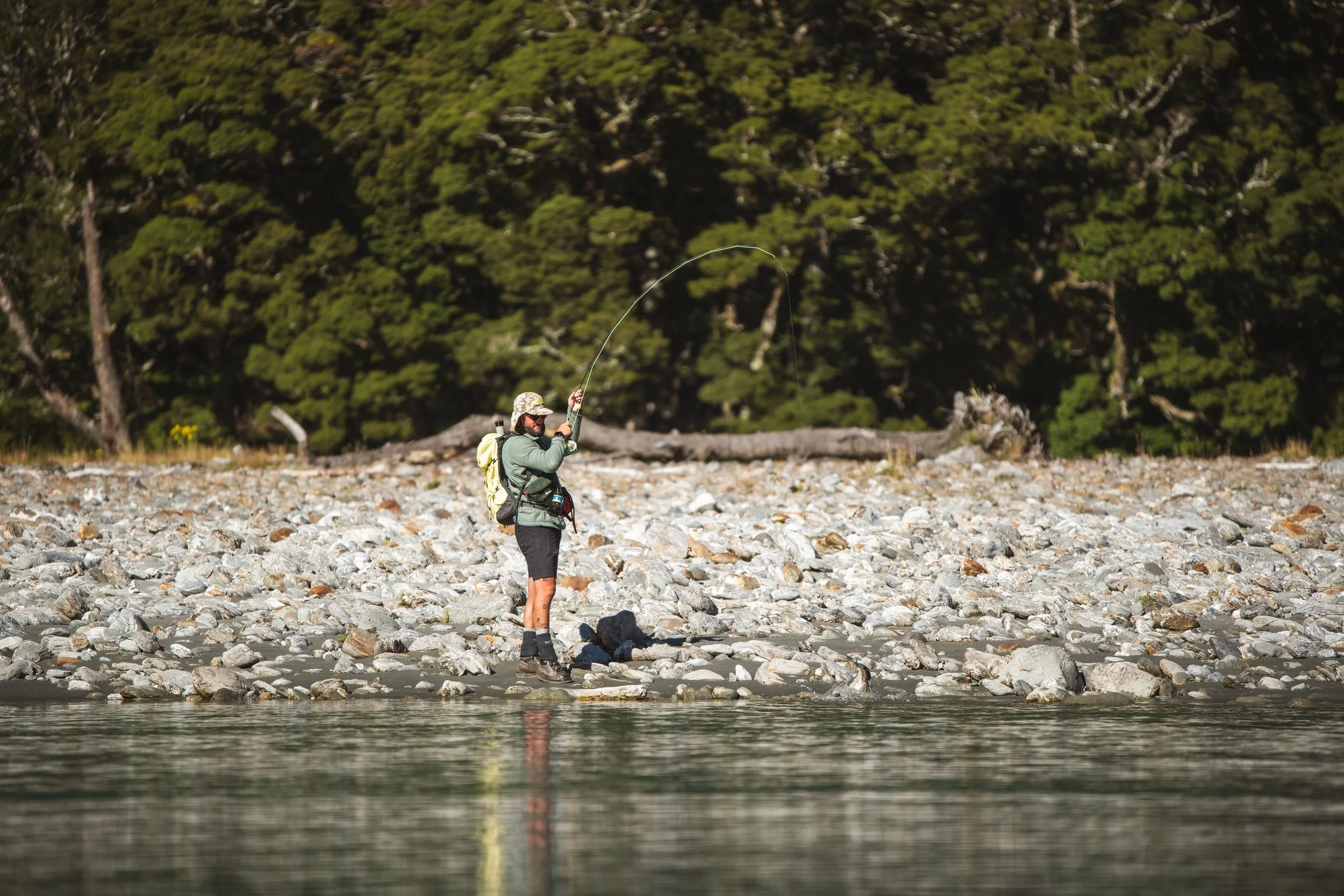 A man standing on a rocky riverbank fly fishing in the river, with a forested hillside in the background.