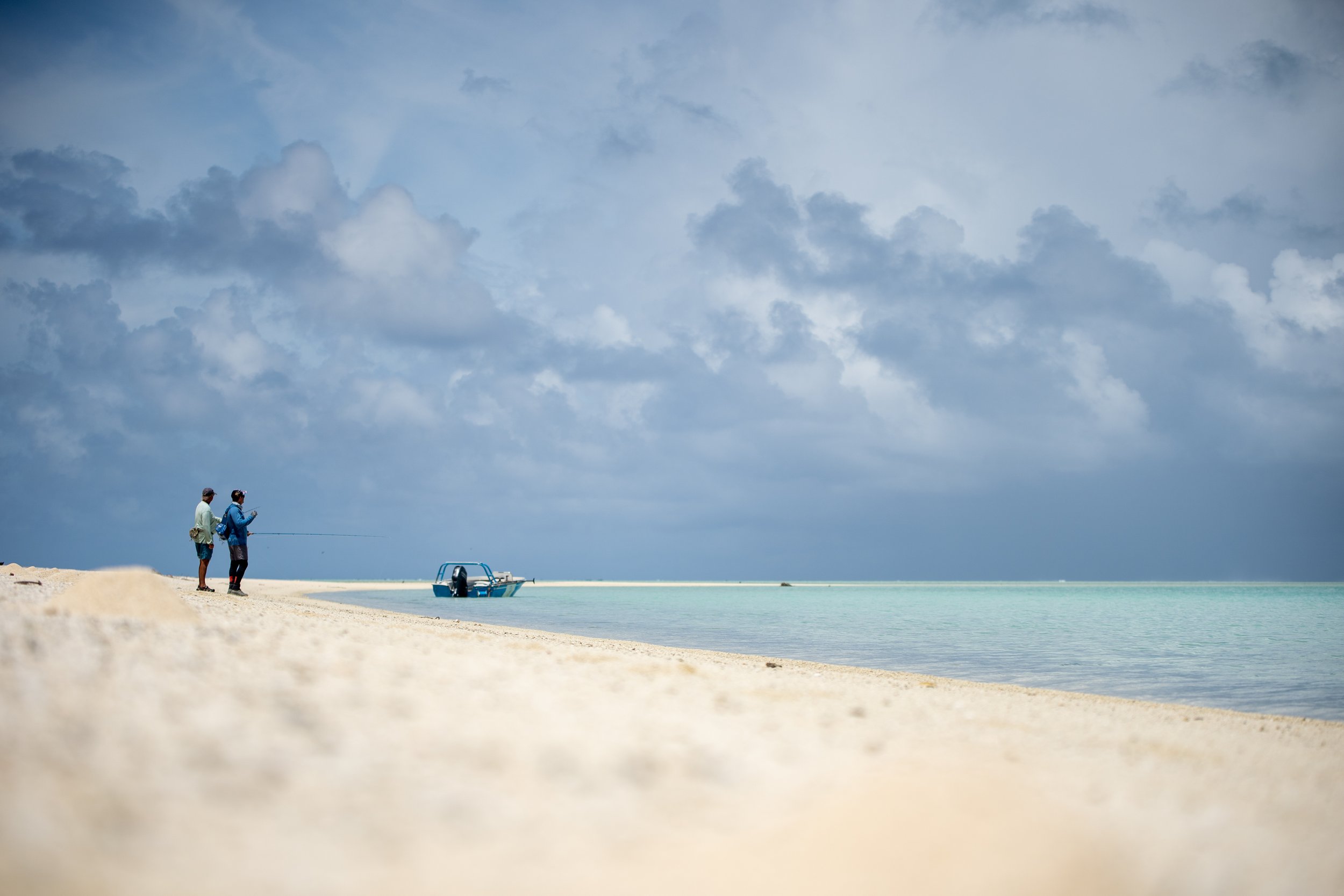 Two people fishing on a sandy beach under a cloudy sky, with a small boat anchored close to the shore.