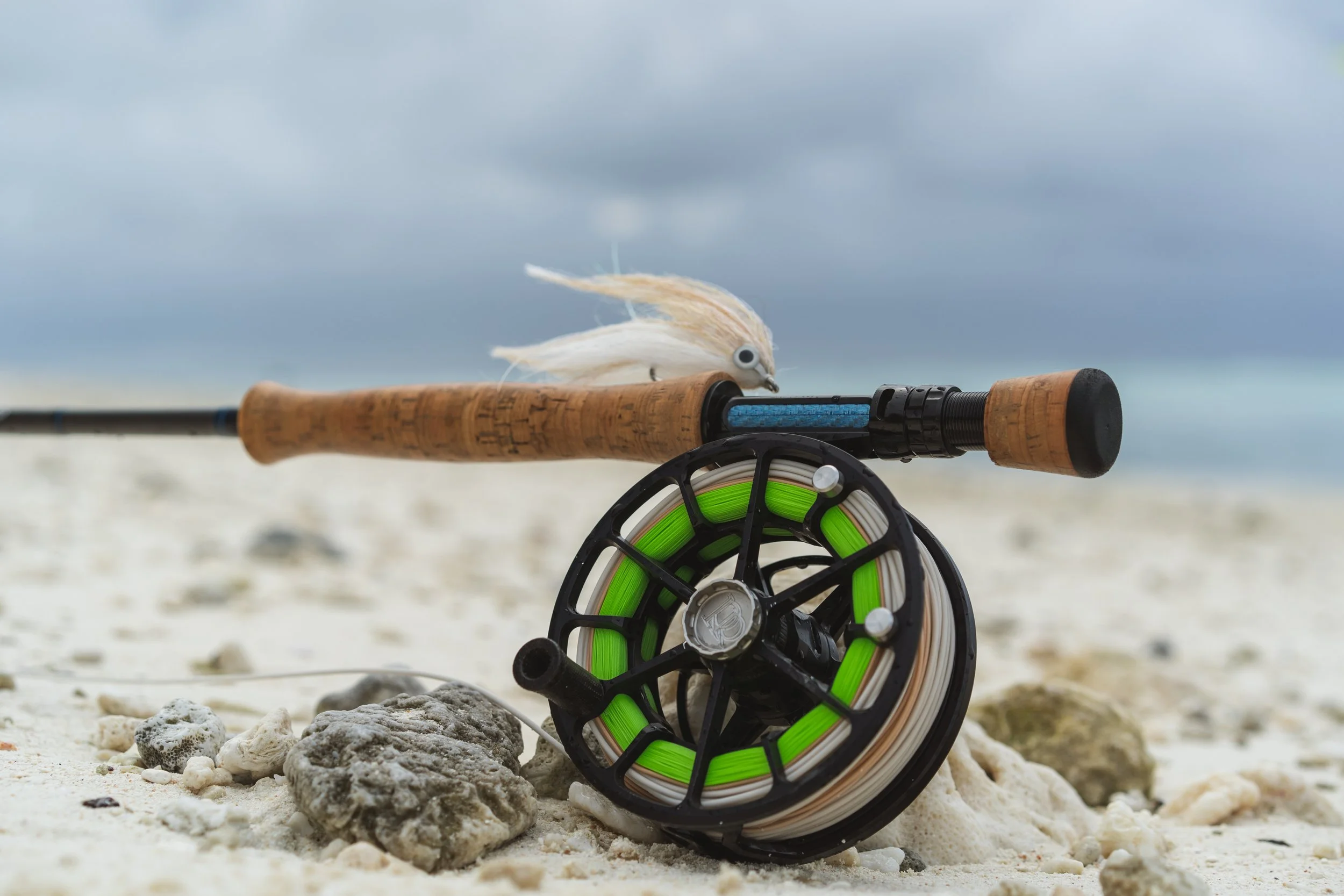 A fishing rod with a white baitfish lure attached, resting on the sandy beach near rocks under a cloudy sky.