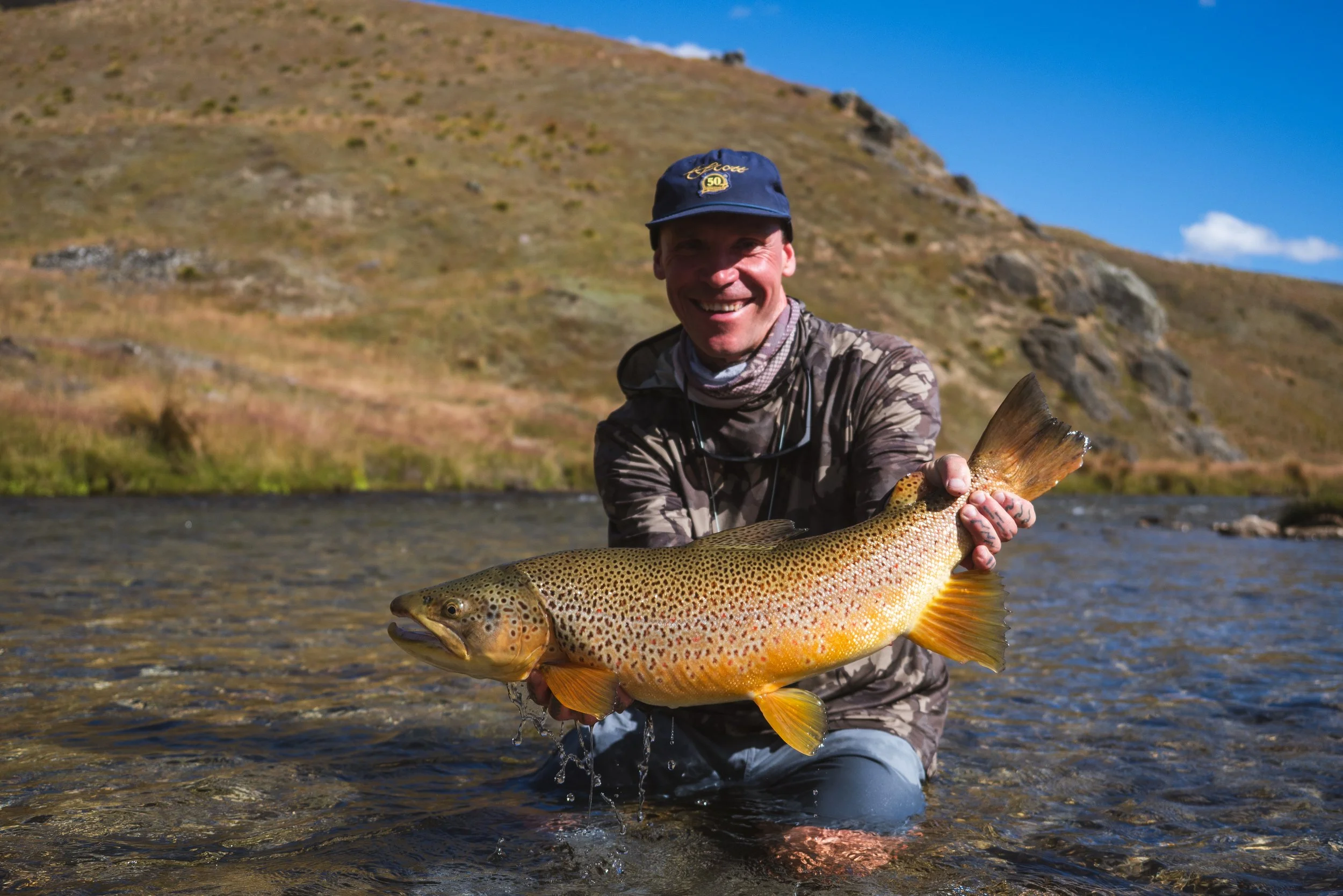 A man smiling while holding a large fish he caught in a river, with hills and clear blue sky in the background.