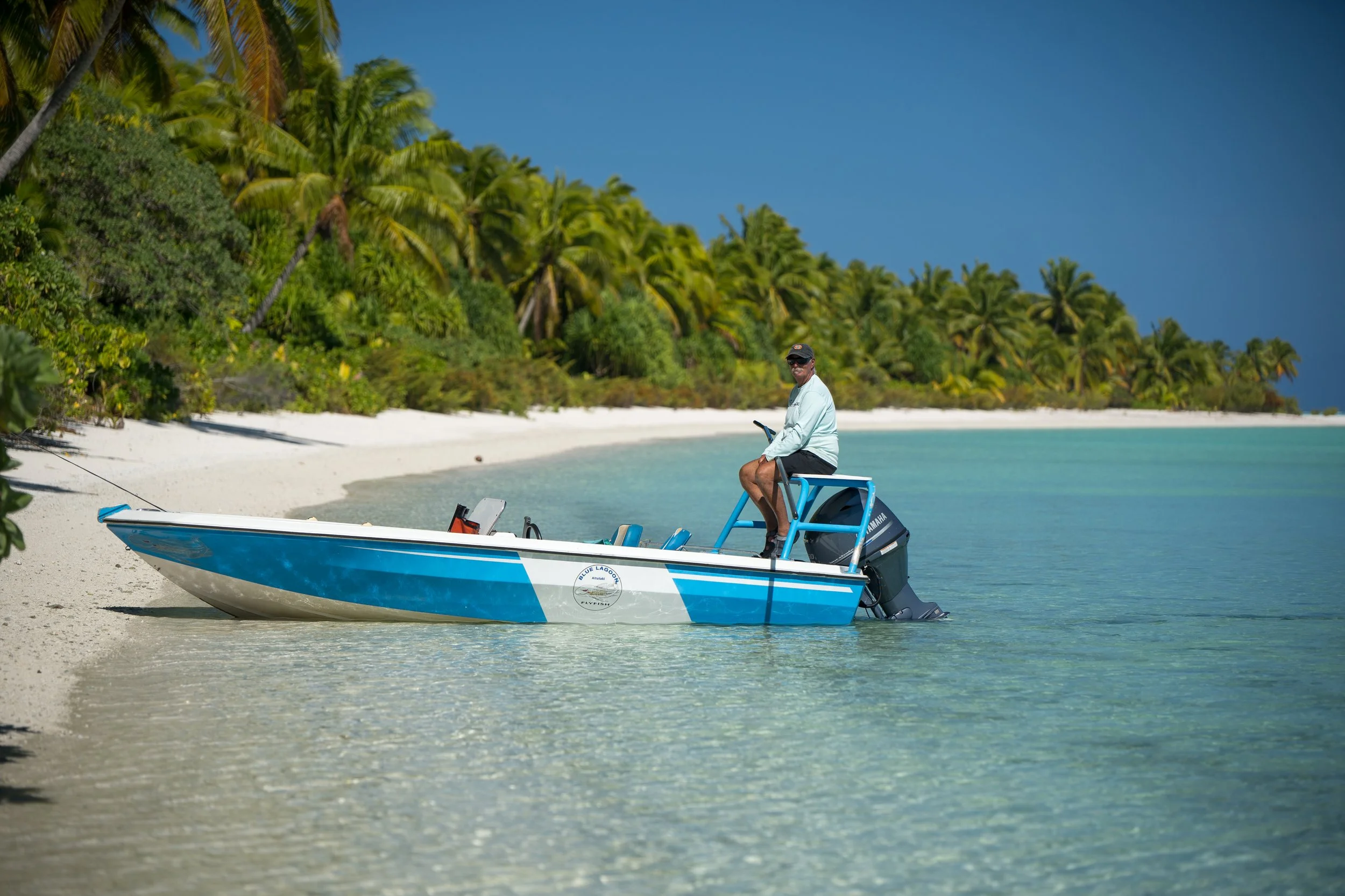 A man sitting on a blue boat with an outboard motor at a sandy beach, with lush green palm trees and a clear blue sky in the background.