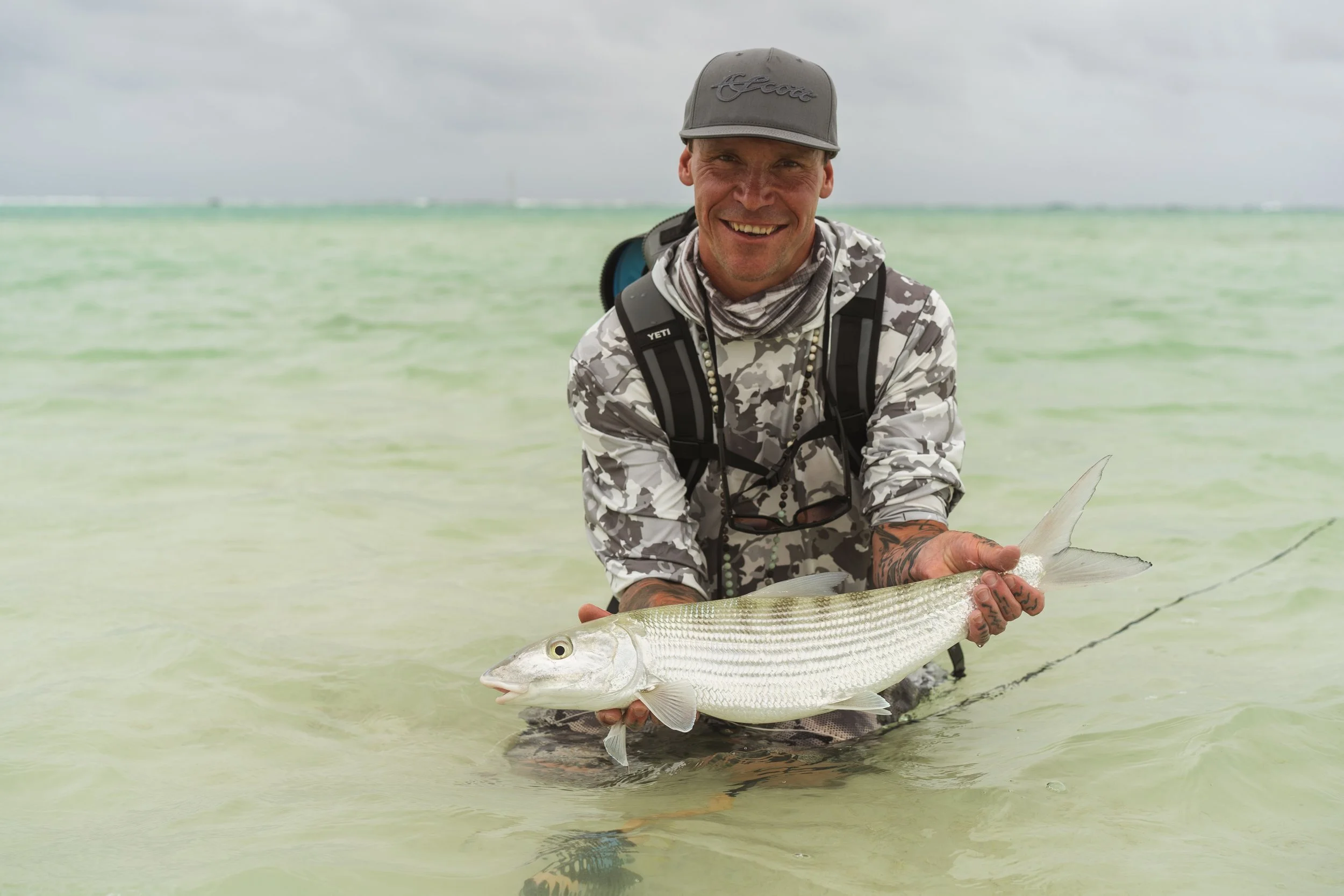 A man in camouflage clothing standing in shallow water, smiling while holding a large fish.