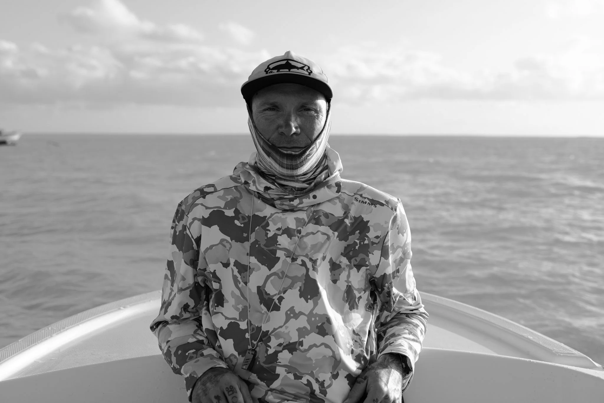 A man wearing a cap, scarf, and camouflage jacket sits on a boat with a body of water and a partly cloudy sky in the background.