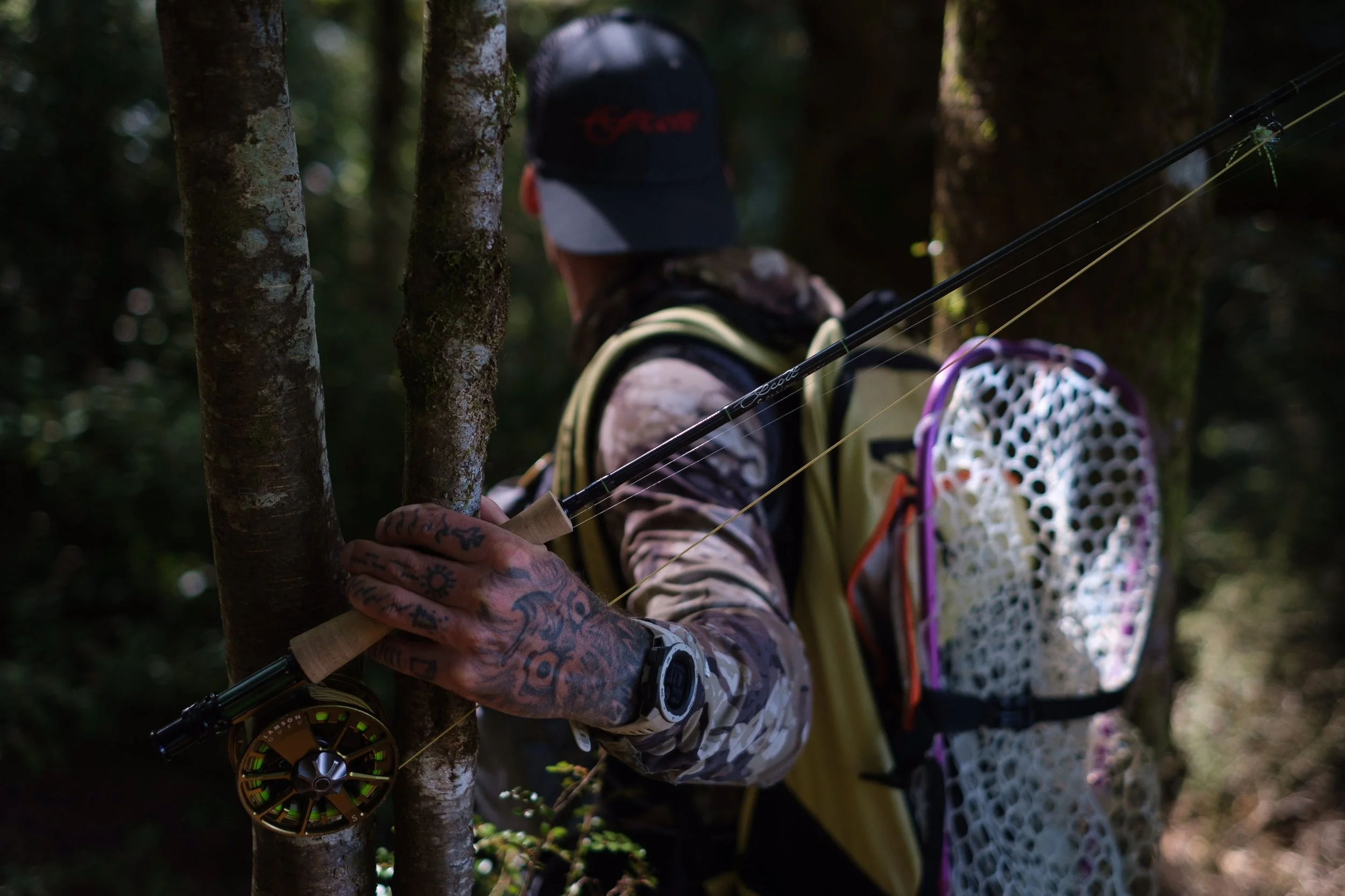 Person wearing camouflage clothing, a black cap, and a watch, with tattoos on their hand, holding a fishing rod while standing behind trees in a forested area.