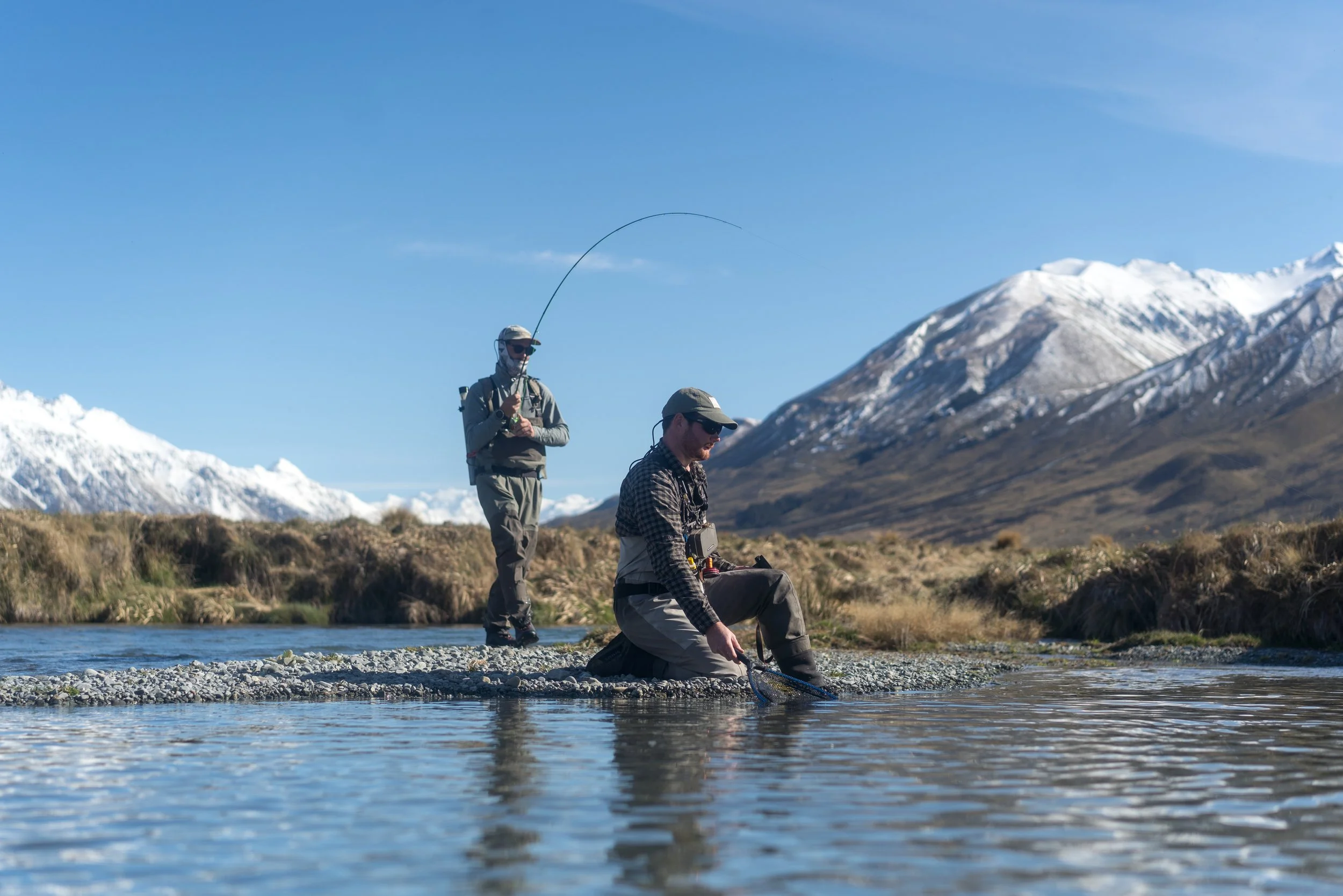 Two men fishing in a mountain stream with snow-capped peaks in the background, one standing and casting a fishing line, the other crouching and preparing to fish.