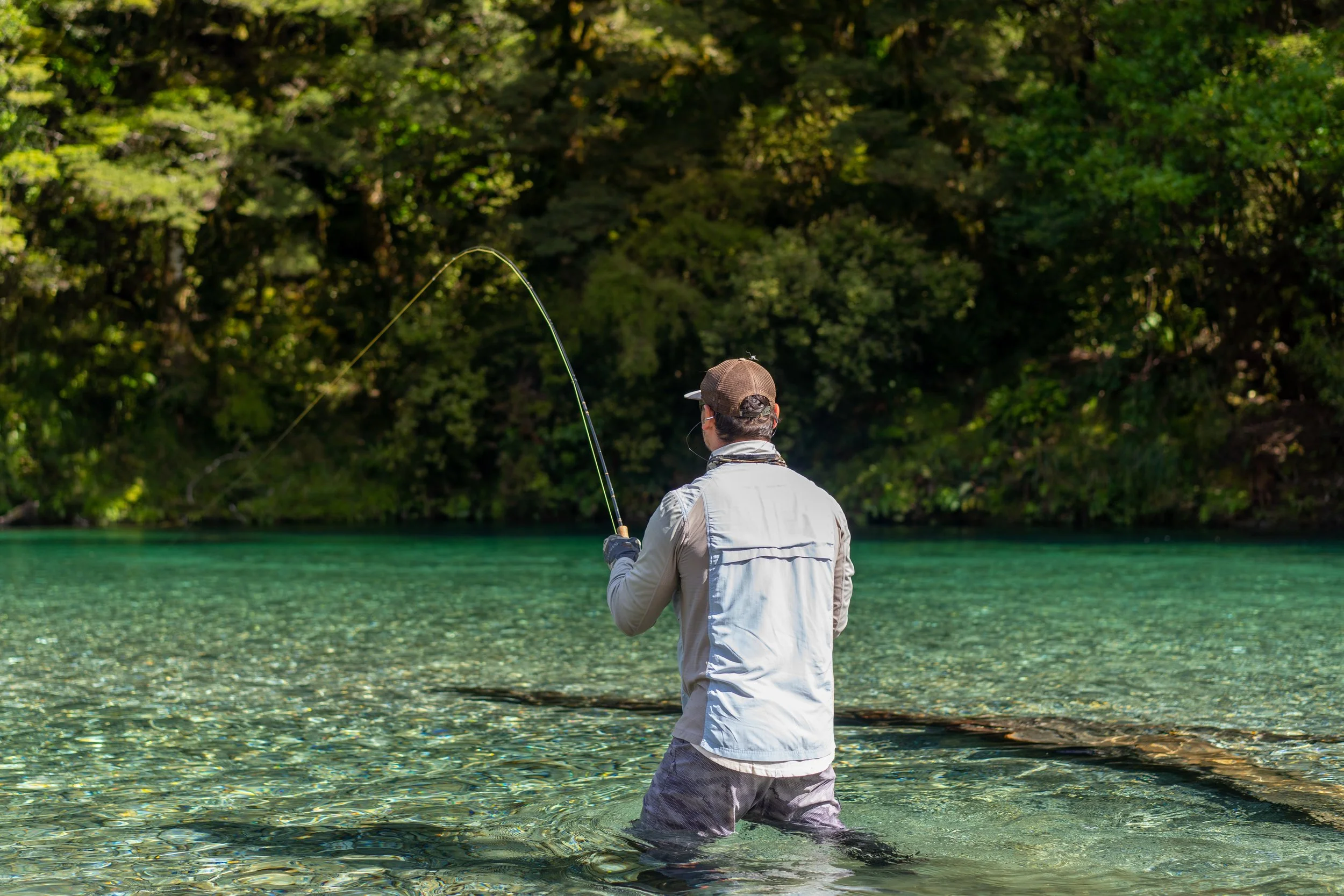 A man fishing in a clear green lake with dense trees in the background.