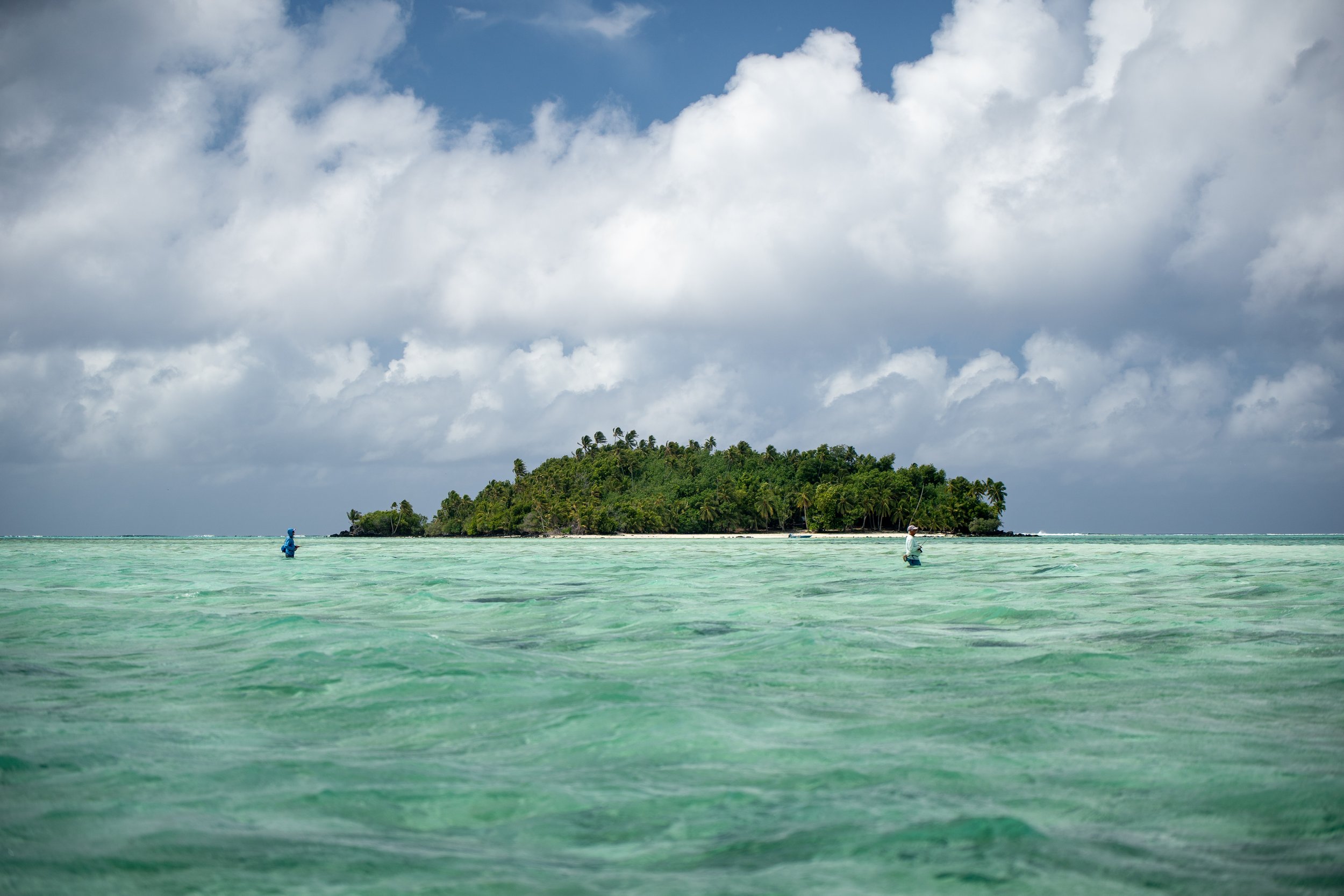 A tropical island surrounded by clear turquoise waters with two people fishing in the foreground, under a partly cloudy sky.