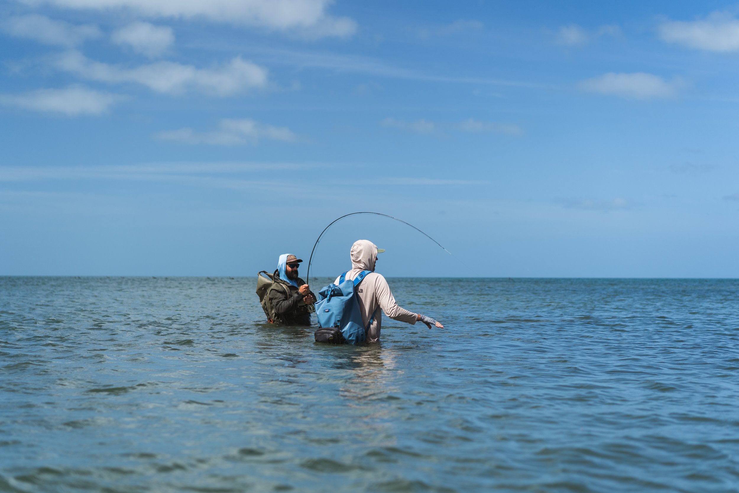 Two people standing waist-deep in water at the beach, one holding a fishing rod, under a partly cloudy blue sky.