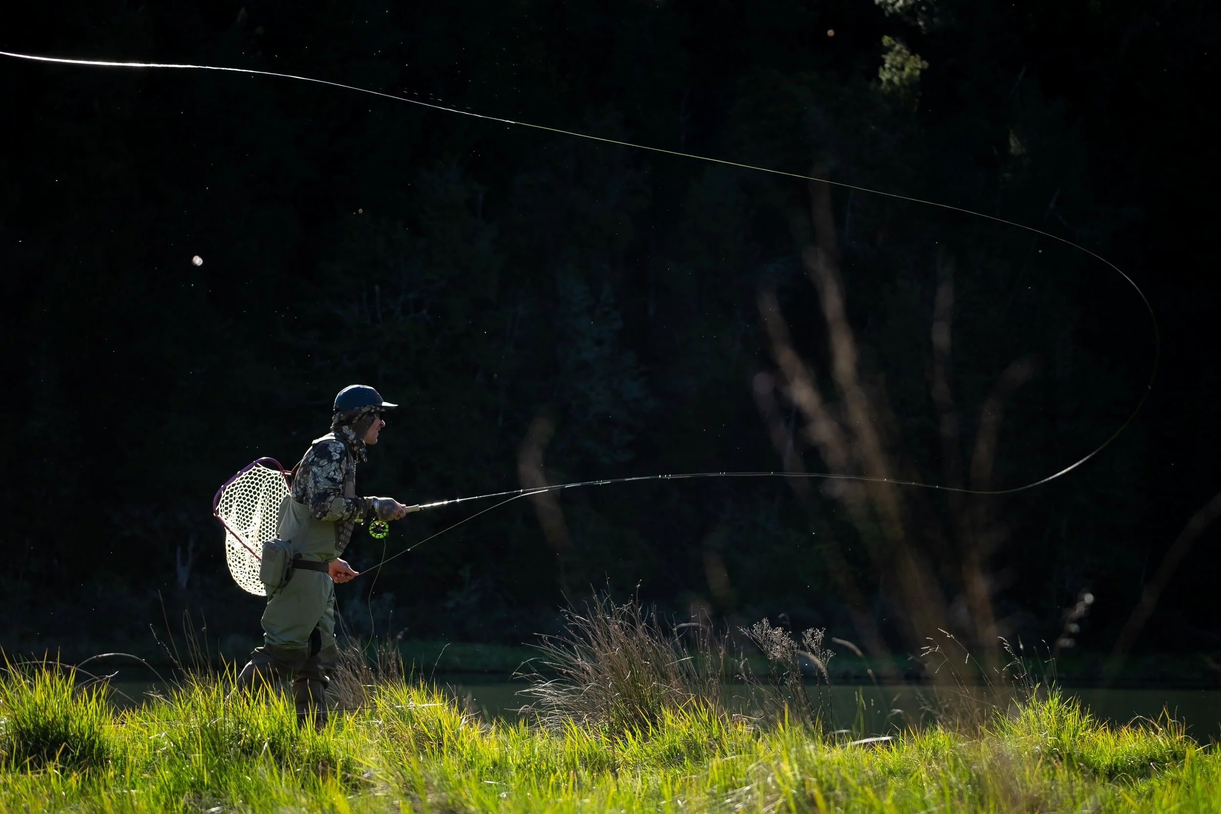 A person fishing at night by a river, illuminated by natural light, with a dark forest background and starry sky visible.