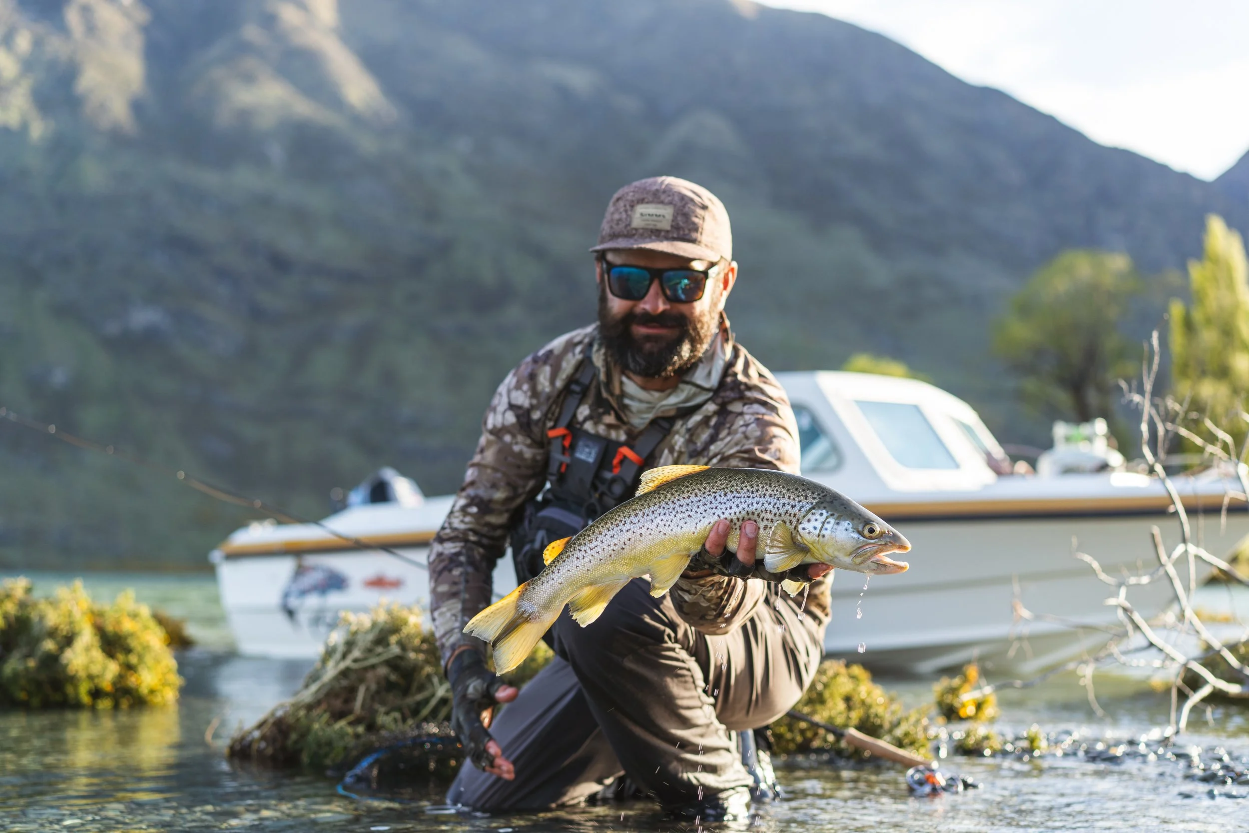 A man wearing camouflage clothing, a cap, and sunglasses kneels in water, holding a large fish with a yellow stripe, with mountains and boats in the background.