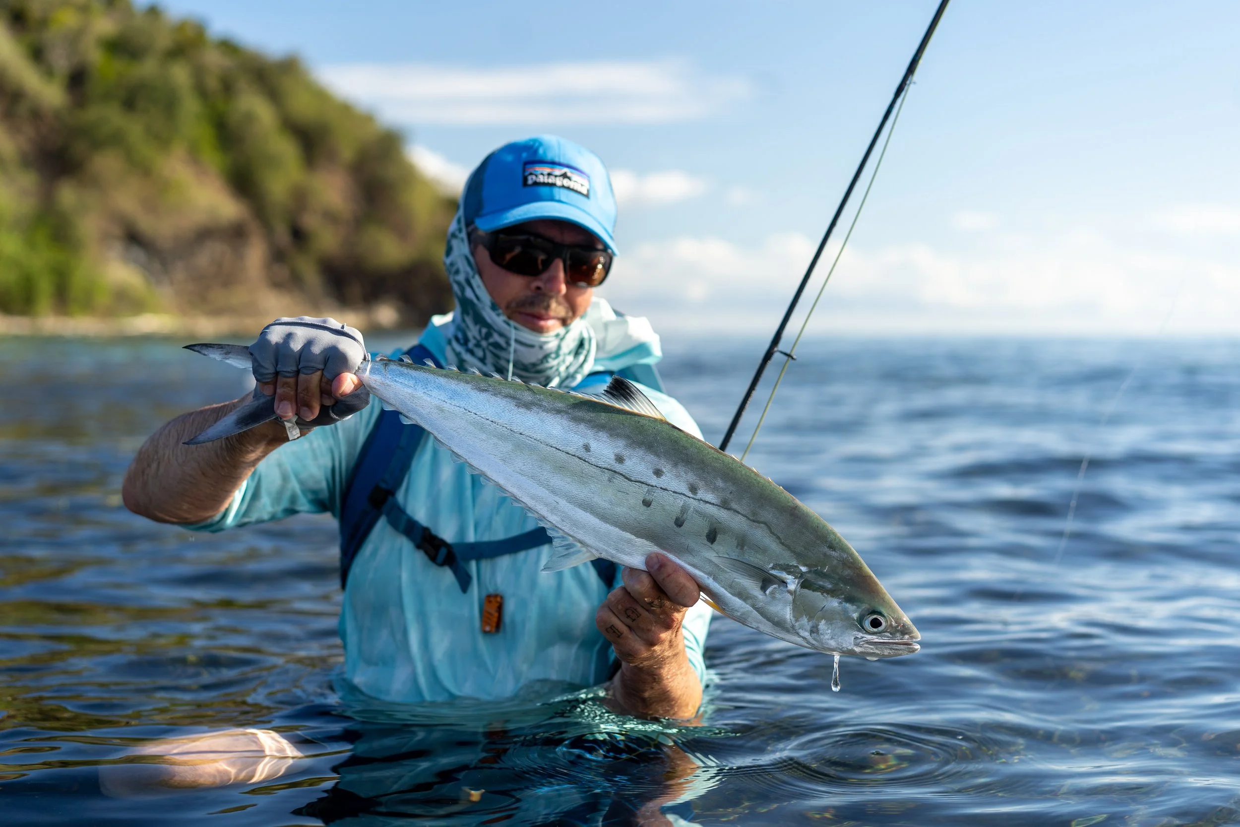 Man in blue hat and sunglasses holding a fishing rod and a fish he caught over water with a green hill in the background.