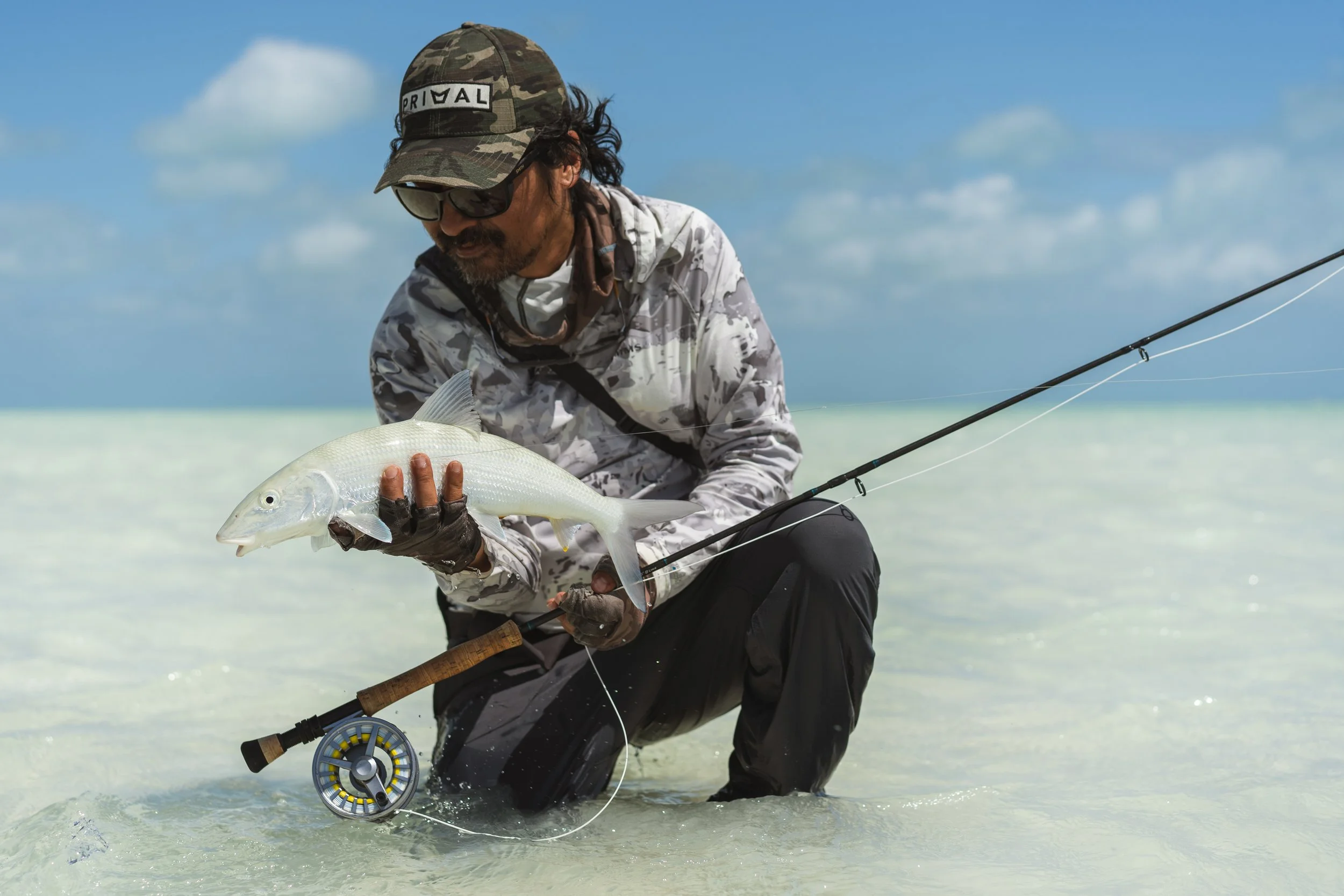 Man in camouflage hat and shirt kneeling in shallow water, holding a fish with a fishing rod nearby, under a partly cloudy sky.