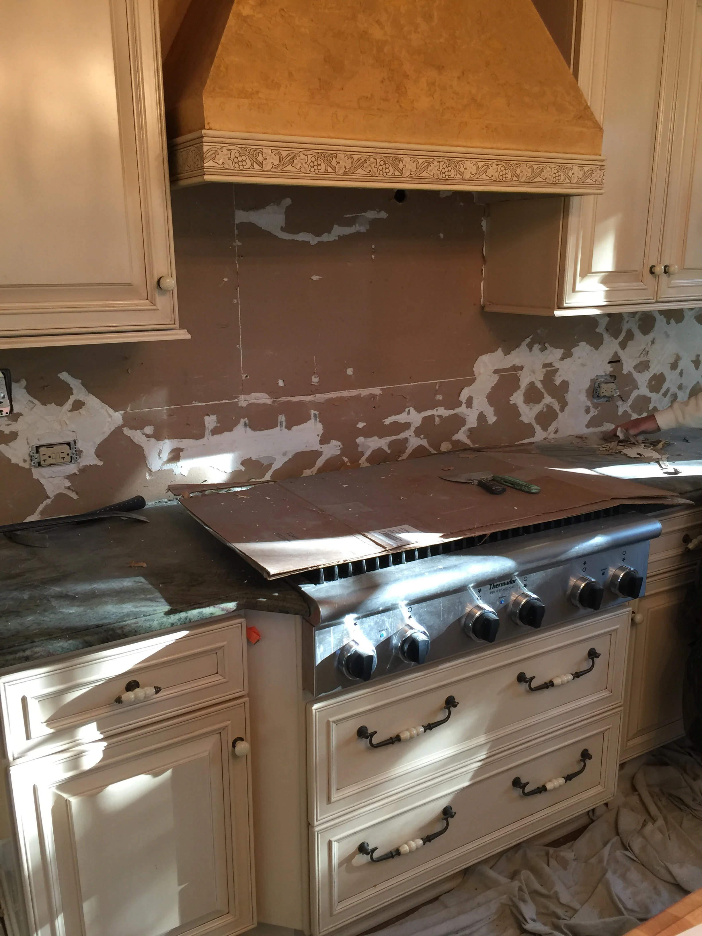 We removed the pastoral backsplash, cabinet hardware, and countertops. The china cabinet found a new home, and the walls were skim-coated smooth for paint. Cream colored cabinets were targeted for new paint. What to do about the heavily textured mustard-colored range hood?