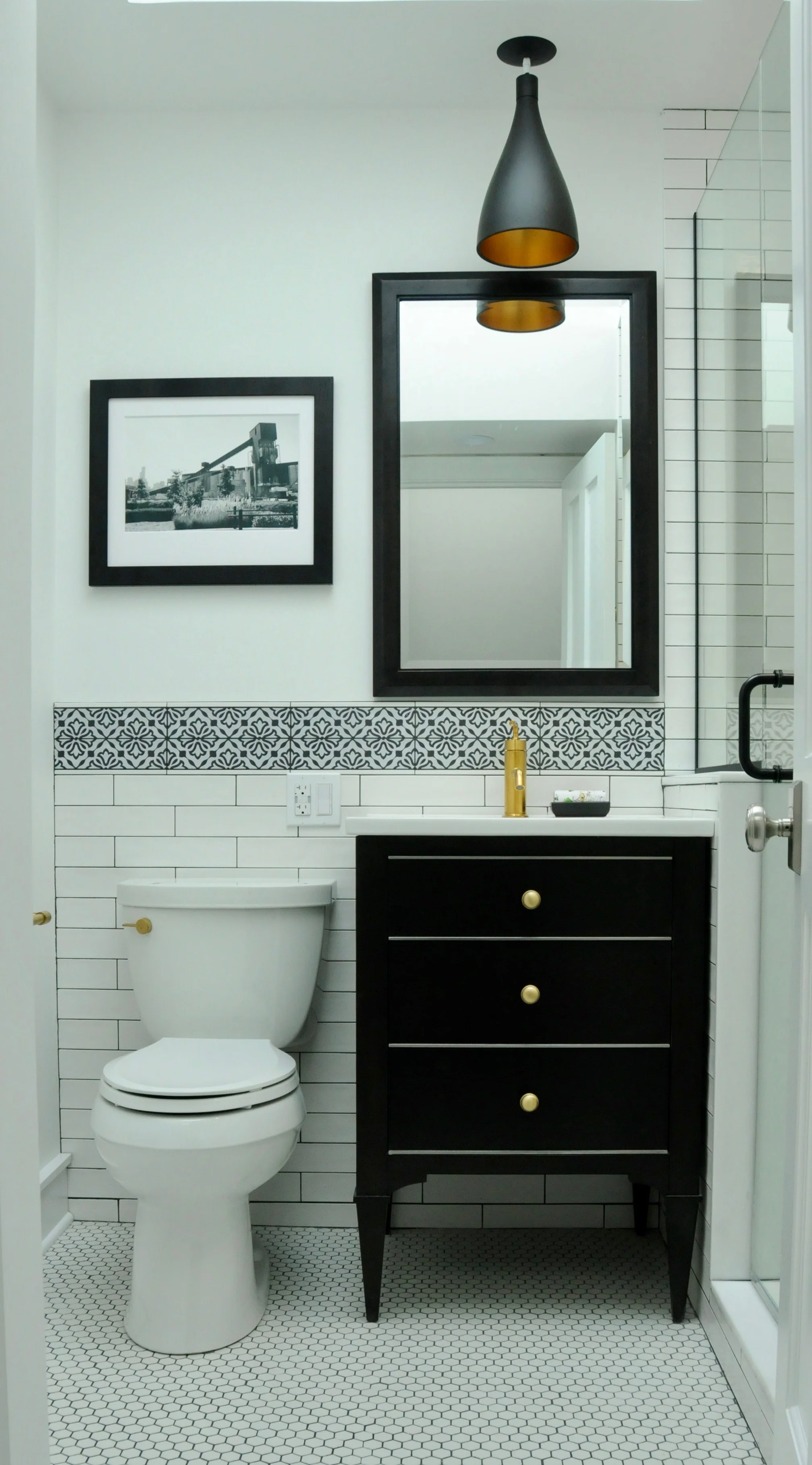 The new bathroom exchanges the short pedestal sink for a full-height black vanity with storage, the polished chrome for warmer tones, and the ongoing maintenance of white grout for charcoal. 