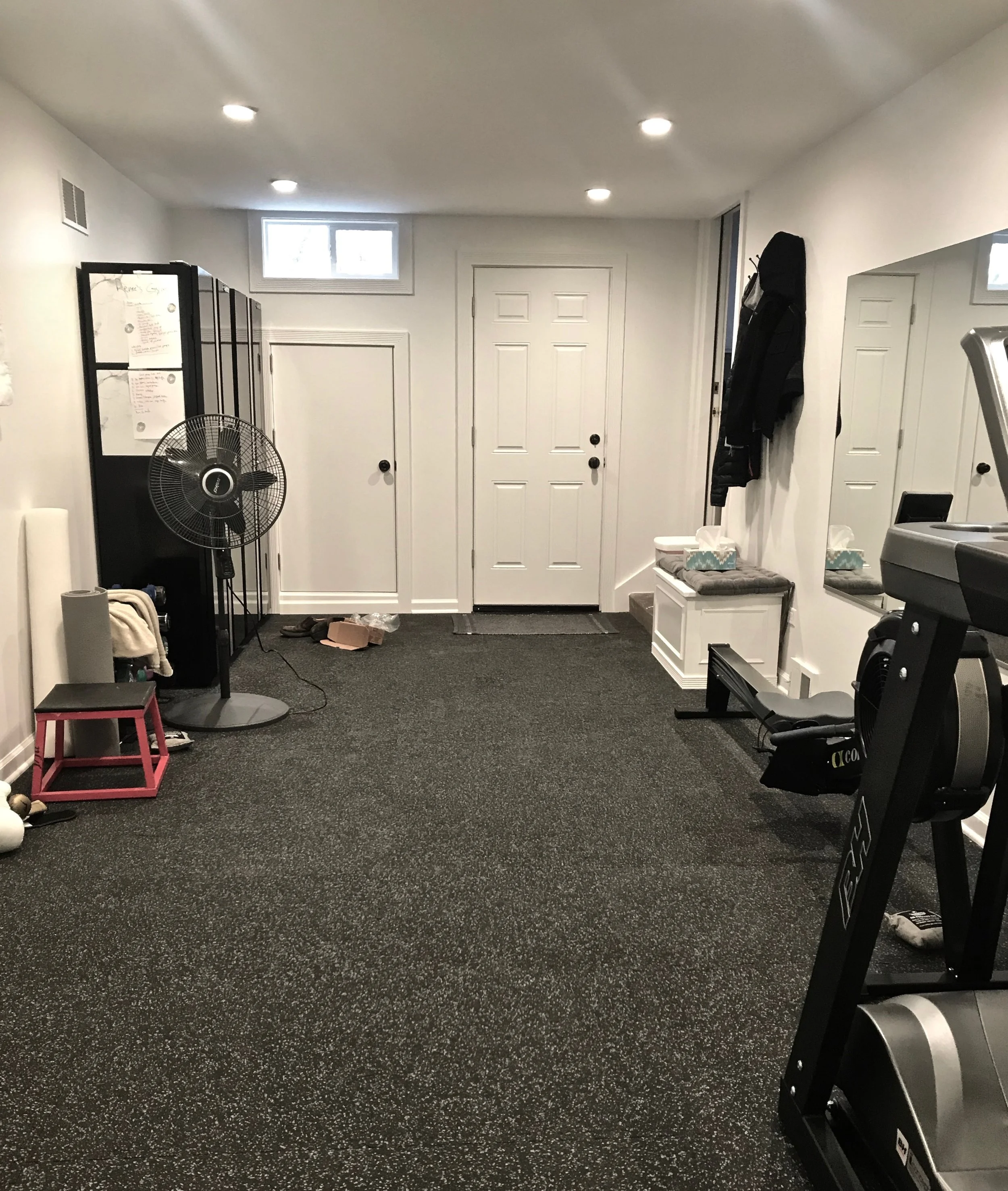 A view of the entry to the gym, with closed storage, new exterior door, new windows, and lockers. The clients can check their form in the 6’ x 4’ wall mirror, too. Heat is drawn from the main ductwork inside the home, and the walls are coated in a special scuff-resistant paint.