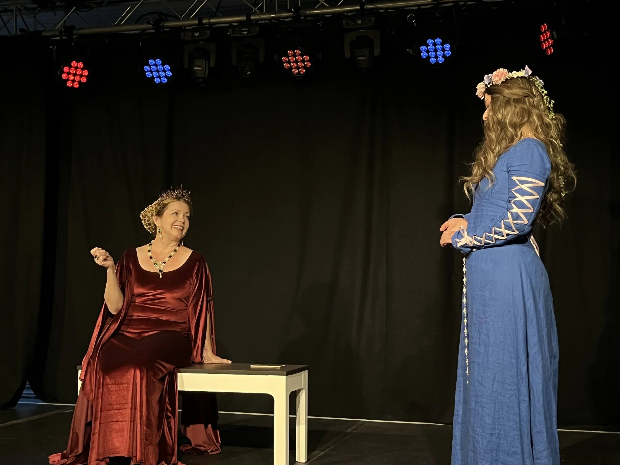 two women medieval dress talking on black stage one sitting on bench