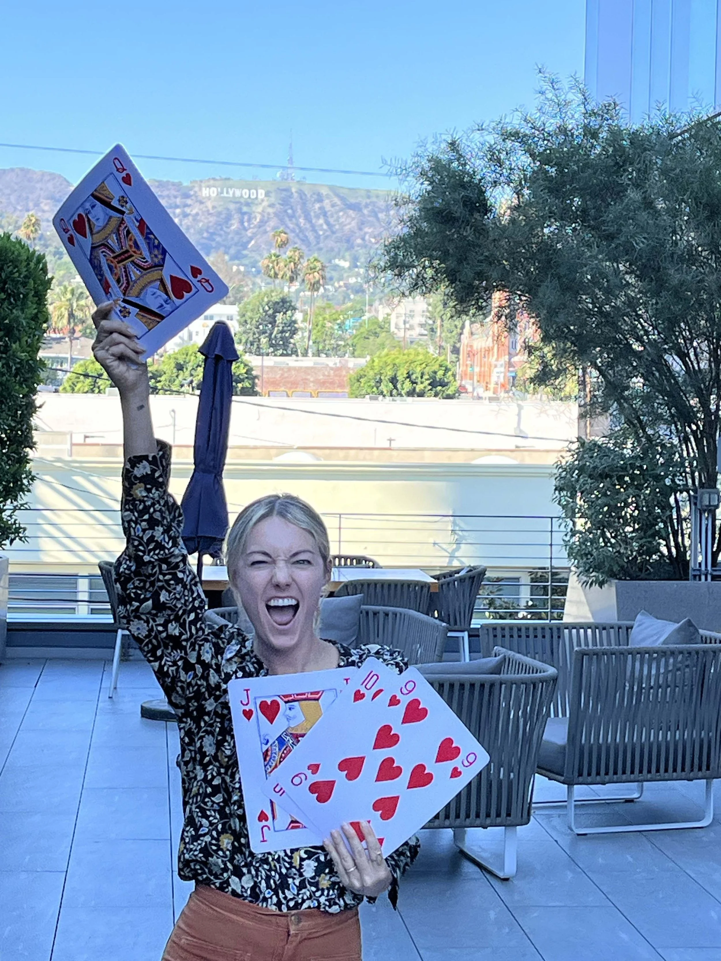 A woman holding oversized playing cards, smiling and posing outdoors with the Hollywood sign in the background.