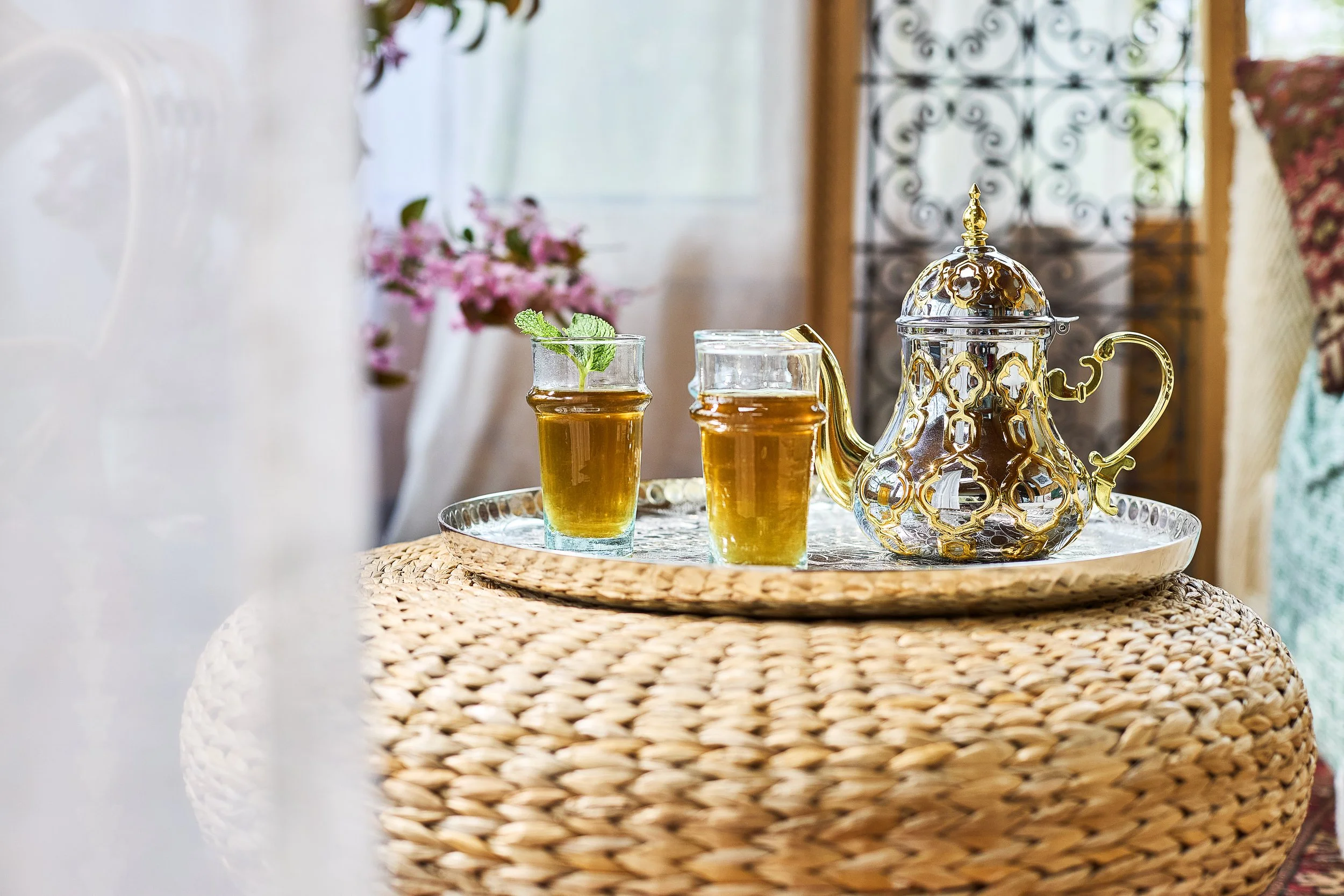 A decorative tea set with a teapot and two glasses of tea on a woven round table, with a blurred background of a room with window, flowers, and ornate iron grille.