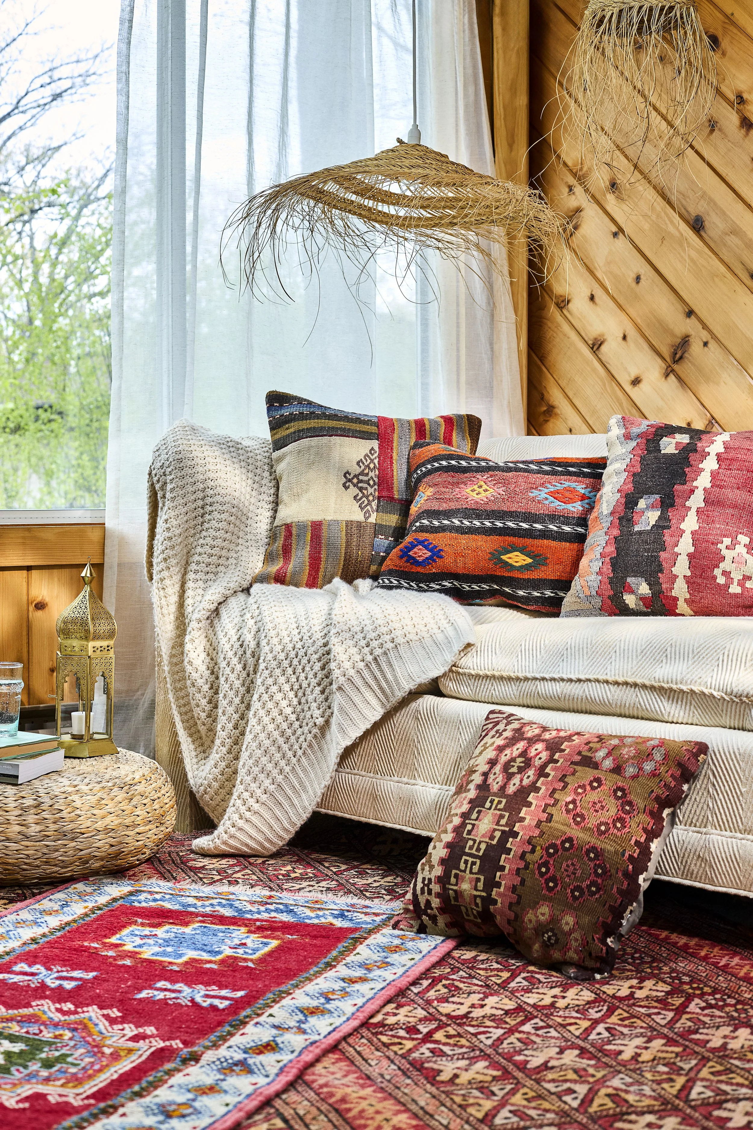 A cozy living room corner with a sofa decorated with colorful patterned cushions, a textured beige blanket, and a round, woven coffee table holding books and a decorative lantern. The room has a wooden paneled wall, a large window with sheer white cu