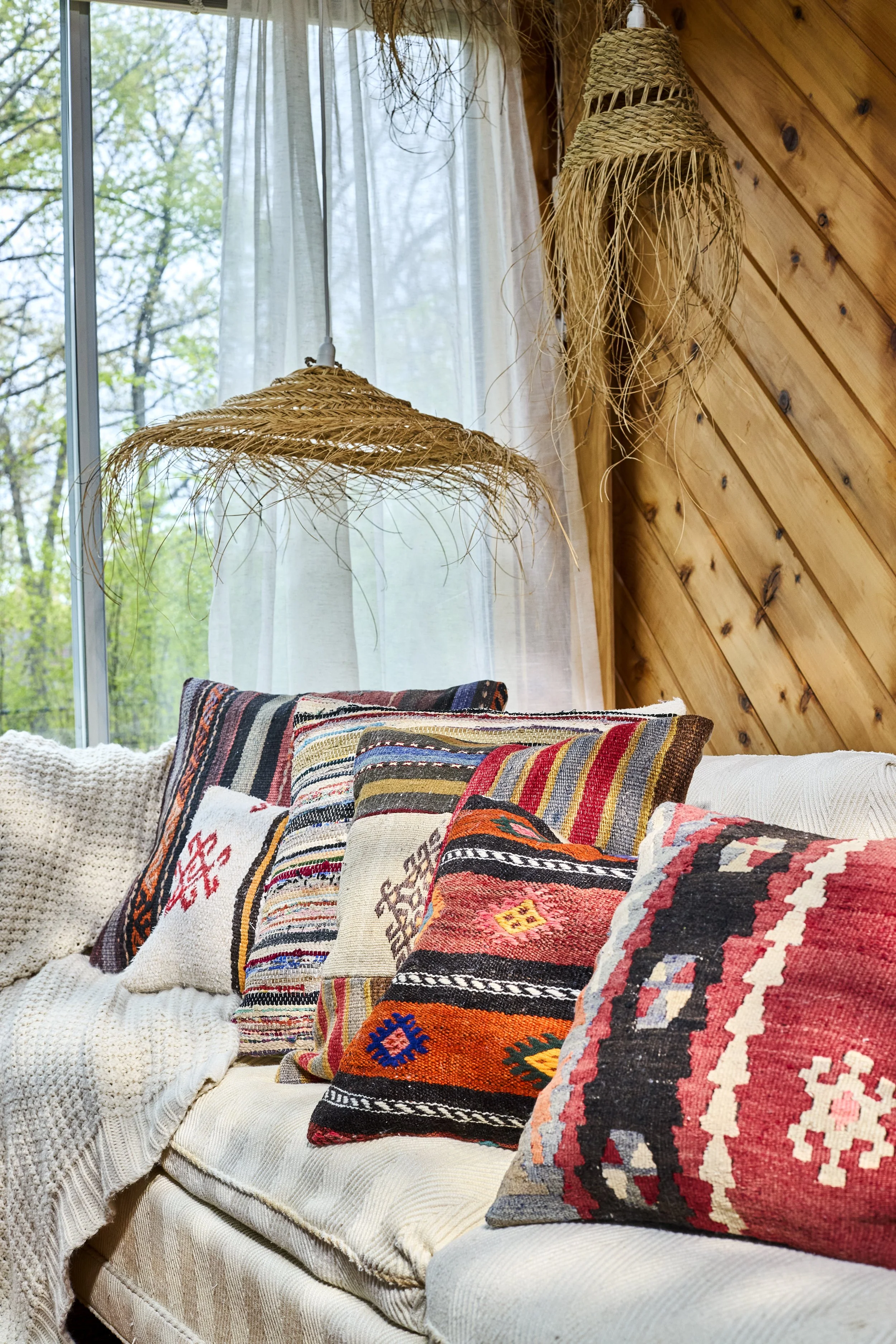 A cozy interior corner with a white textured sofa adorned with colorful, patterned throw pillows, a woven lampshade hanging from the ceiling, a window with sheer curtains, and a wooden-paneled wall.