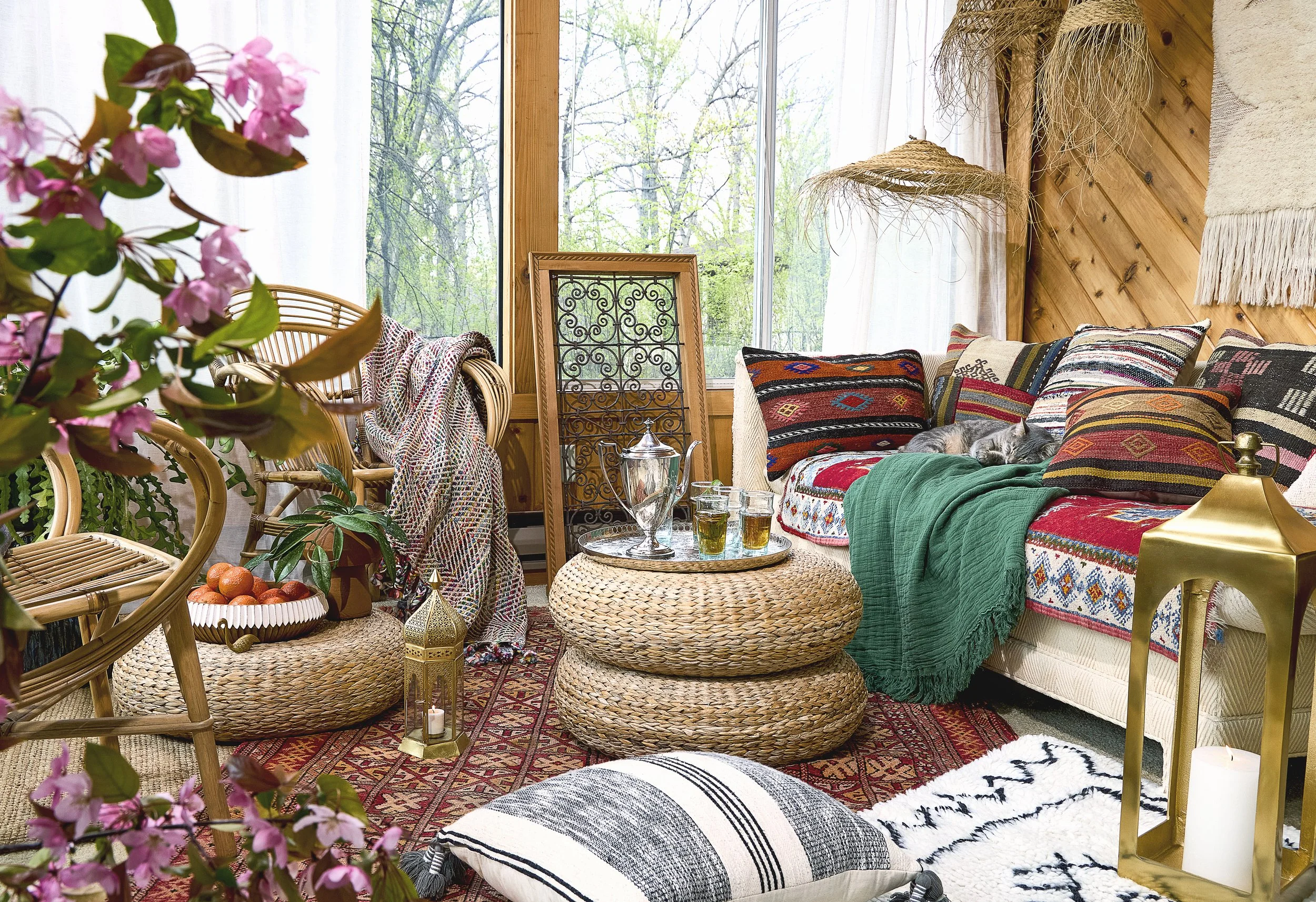 A cozy living room with large windows showcasing a view of trees outside. The space features wicker furniture, colorful patterned pillows, a woven coffee table, and decorative elements like hanging straw lamps and wall hangings. There are pink flower
