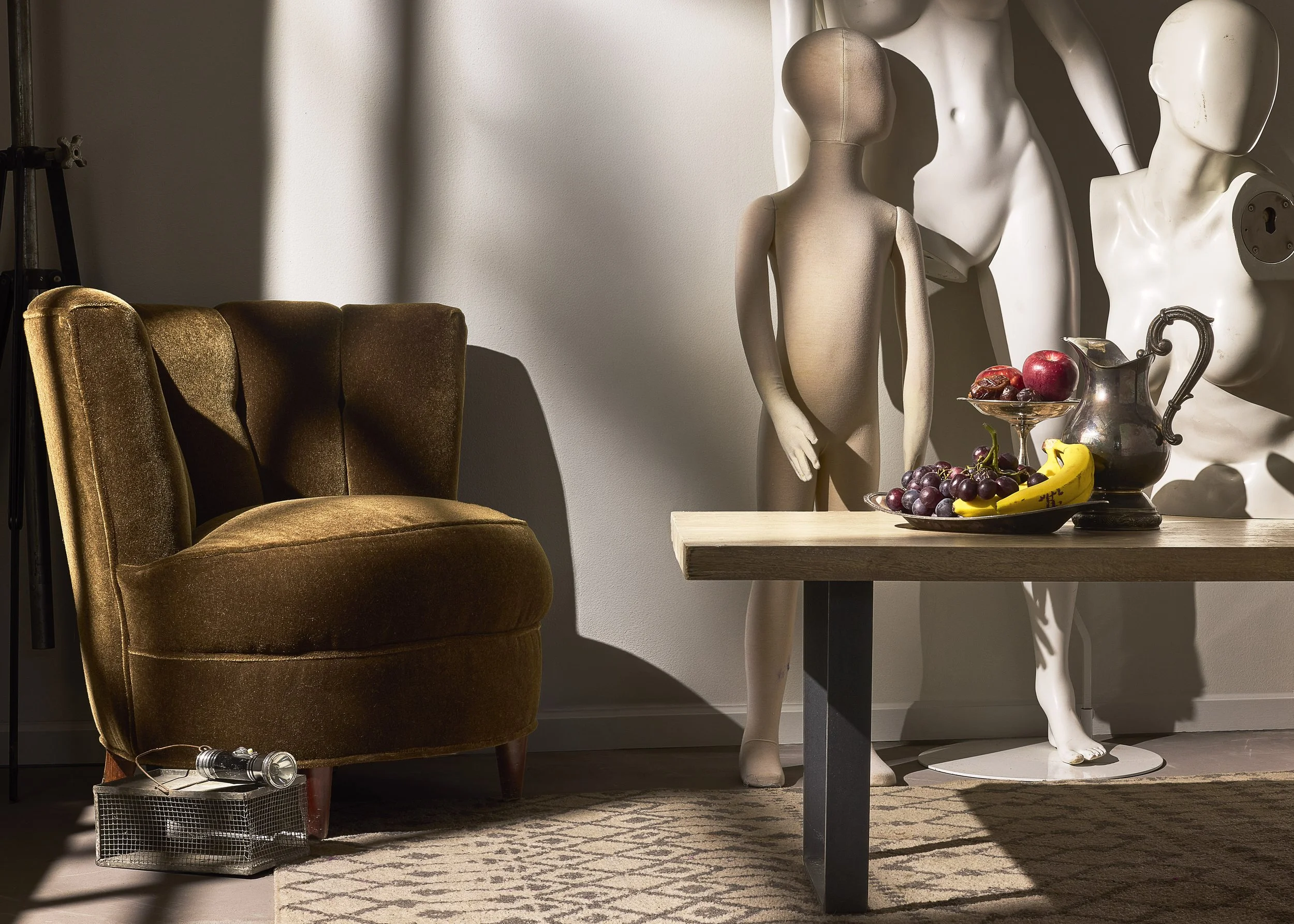 A plush, brown vintage armchair near a light-colored wall, with a flashlight on the floor and a metallic crate beneath it. Next to the chair, there's a rectangular wooden table with a display of assorted fruit including apples, grapes, a banana, and 
