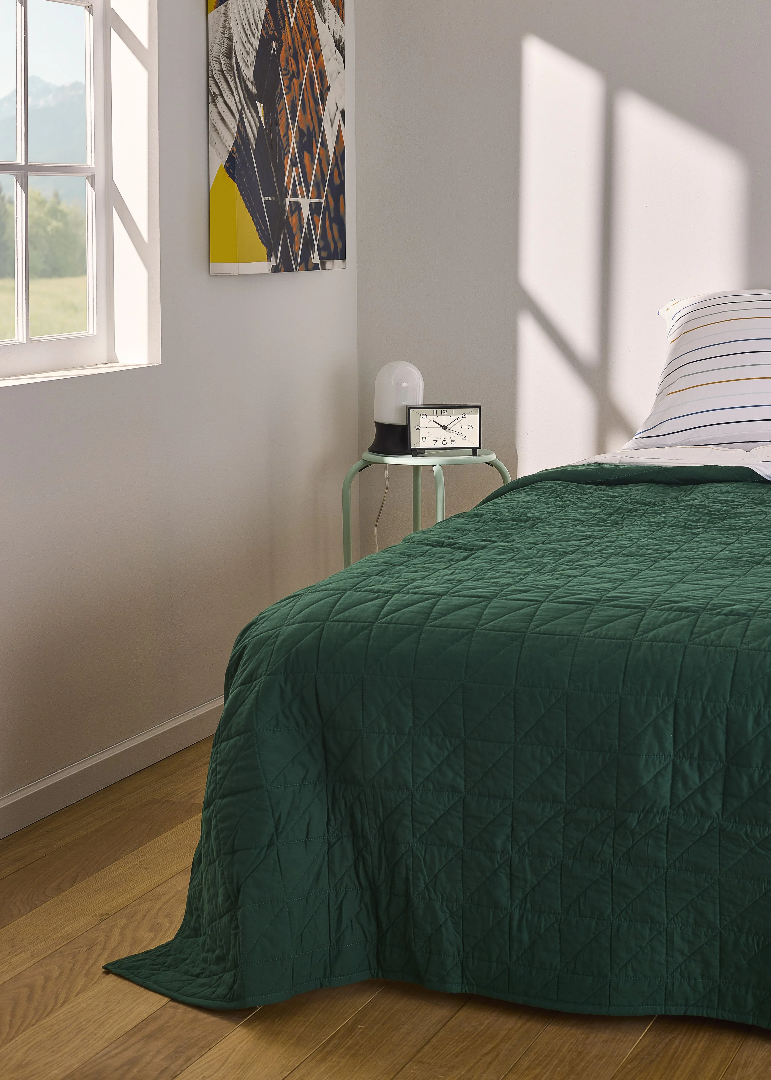 A bedroom scene featuring a bed with a green quilted bedspread and a striped pillow. Next to the bed, there is a small round table holding a white lamp and a black framed clock. There is a window on the left side casting sunlight and shadows on the w