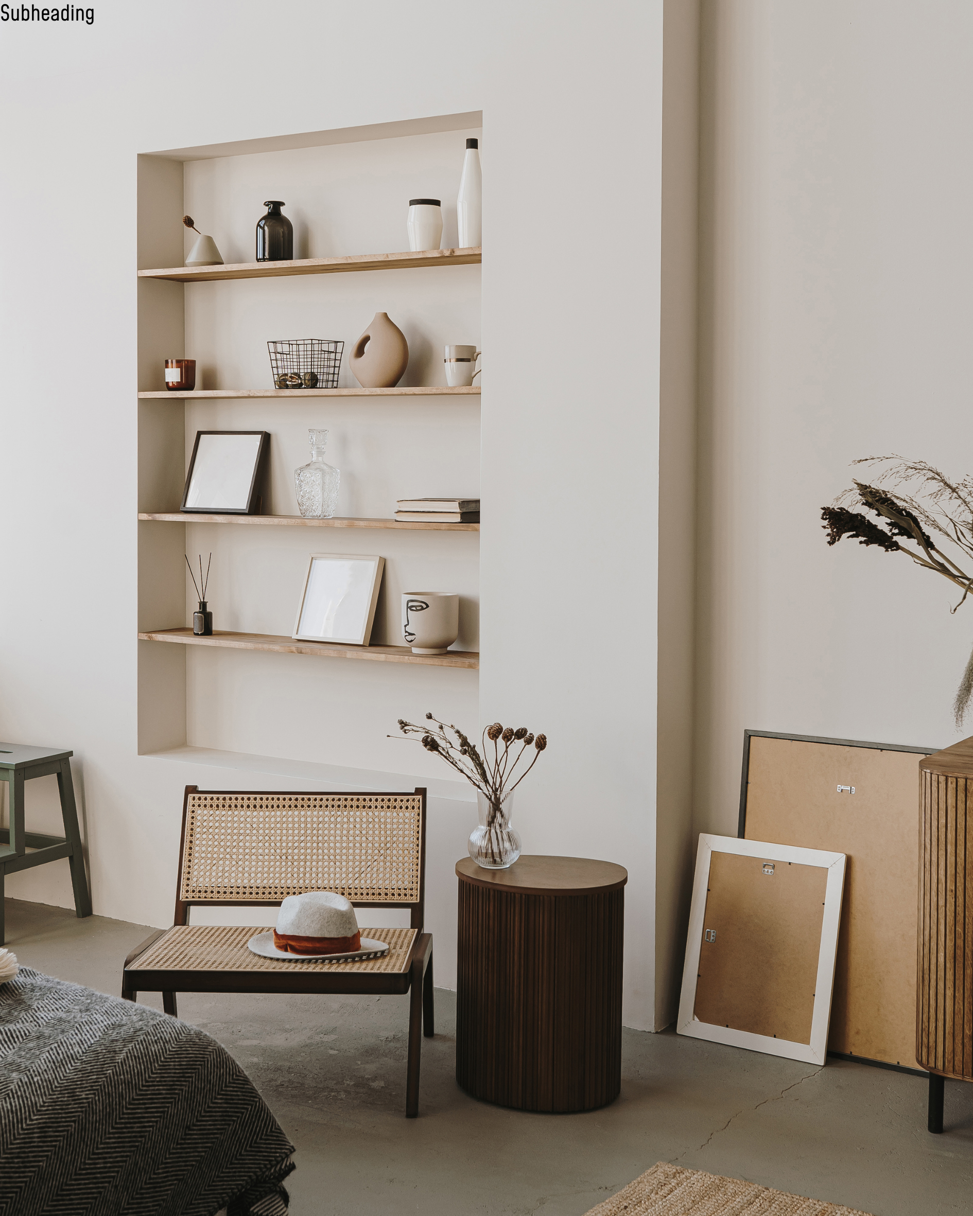 Interior of a modern room with minimalistic decor including a wall niche with shelves holding vases, framed pictures, and decorative objects, a small green stool, a wooden side table with a glass vase and dried flowers, and artwork leaning against the wall.