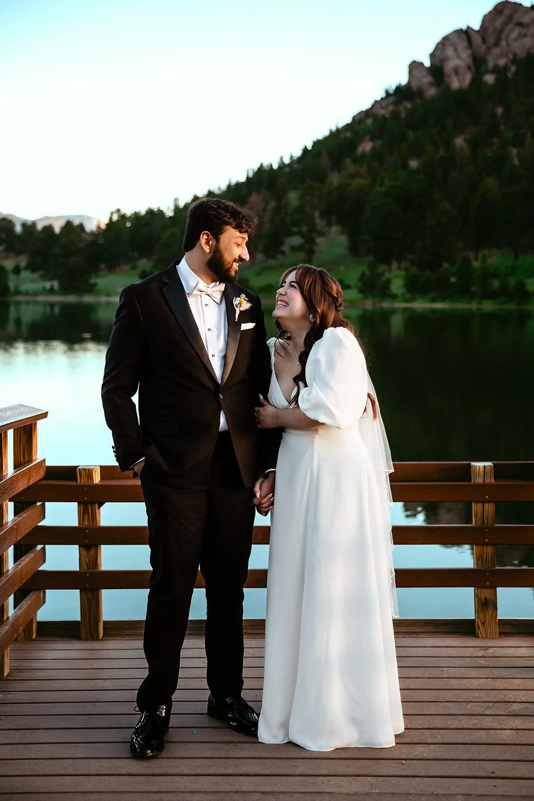 A newlywed couple stands on a wooden pier by a lake, smiling and looking at each other. The groom is dressed in a black tuxedo with a white shirt and bow tie, and the bride is wearing a white wedding dress with puffed sleeves. The background features