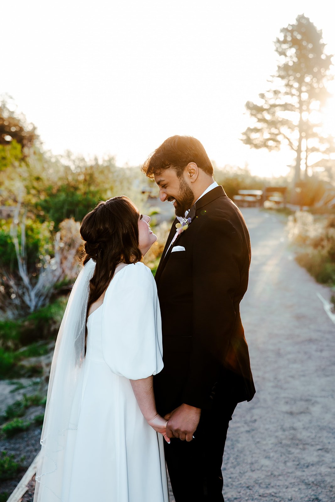 A bride and groom holding hands, smiling, and looking at each other during sunset on a country pathway, with trees and sunlight in the background.