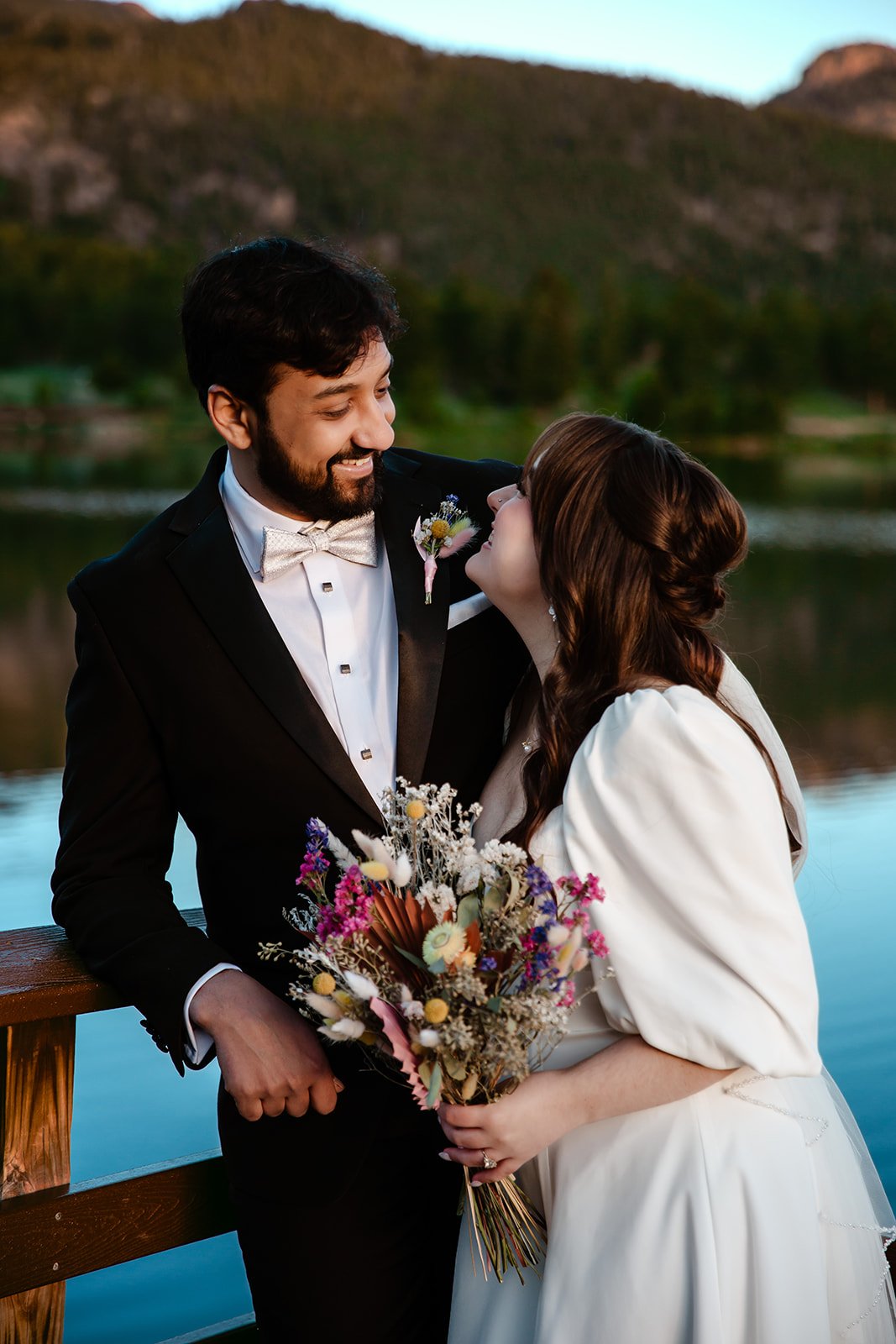 A bride and groom smiling and looking at each other by a lake during sunset, with a mountain in the background.