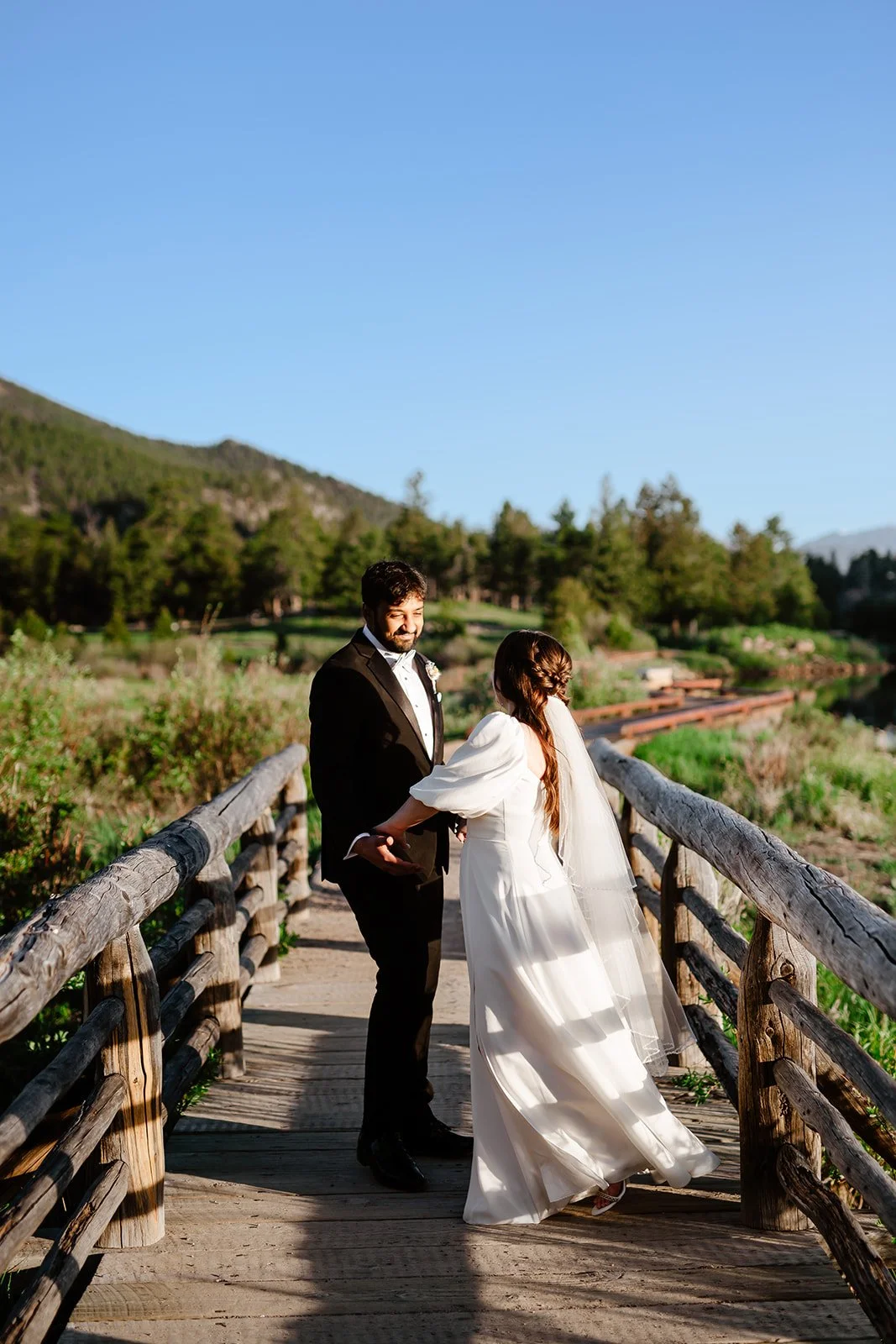 A bride and groom standing on a wooden bridge outdoors during their wedding, holding hands and smiling, with green trees, hills, and blue sky in the background.