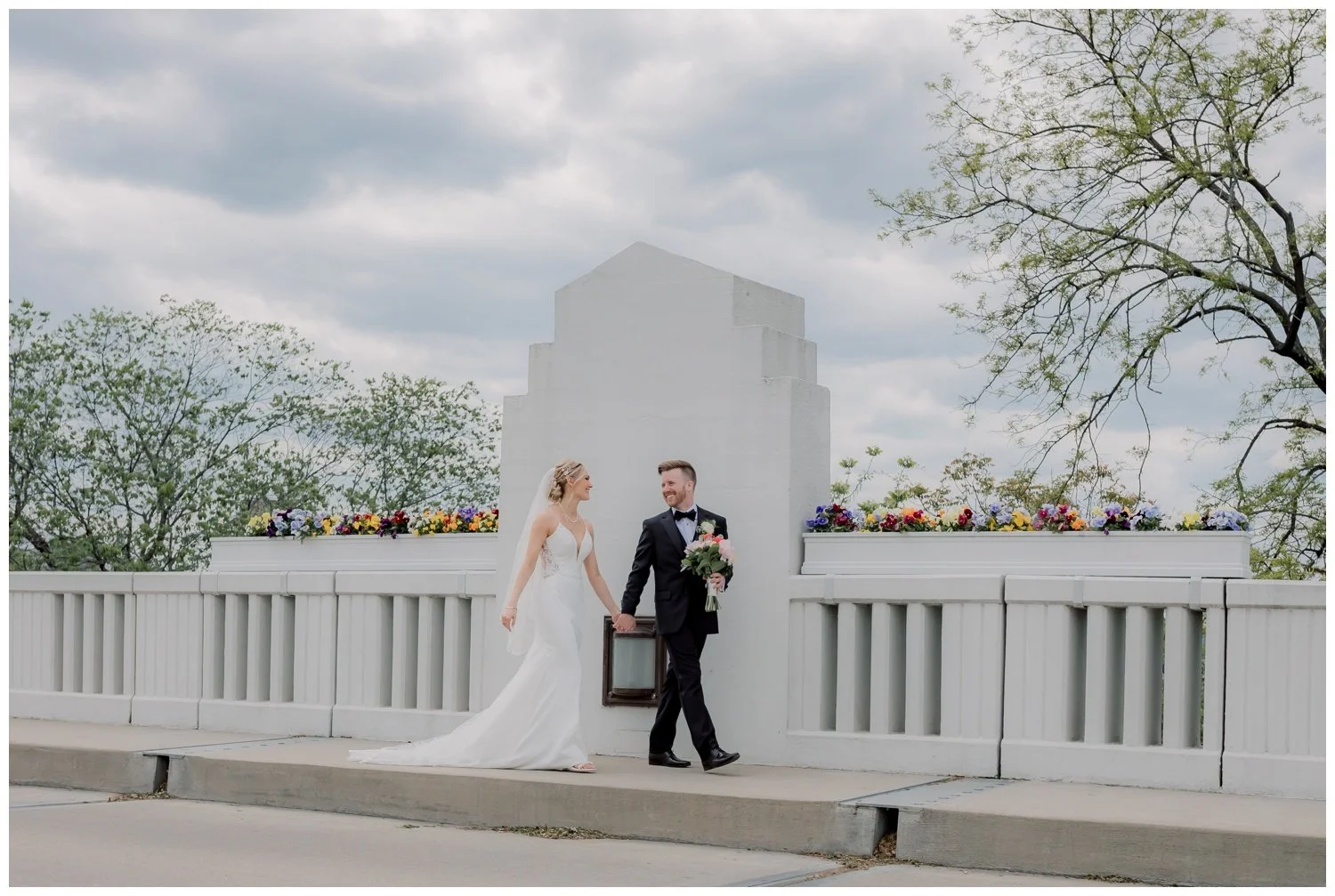 The bride and groom strolling across a white bridge on their wedding day, surrounded by brightly colored flowers.