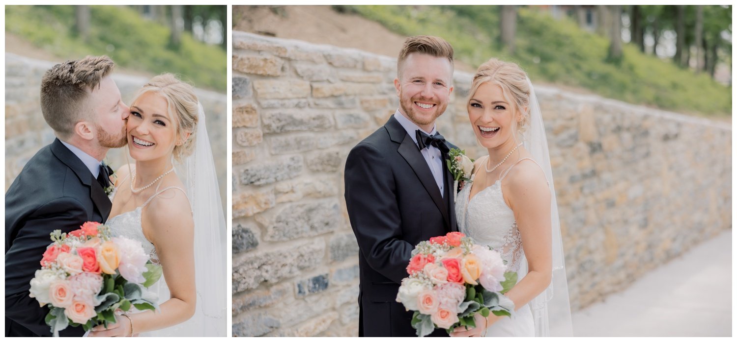 The bride smiling beautifully with her groom kissing her cheek while she holds her brightly colored bouquet in front of a rock wall background.