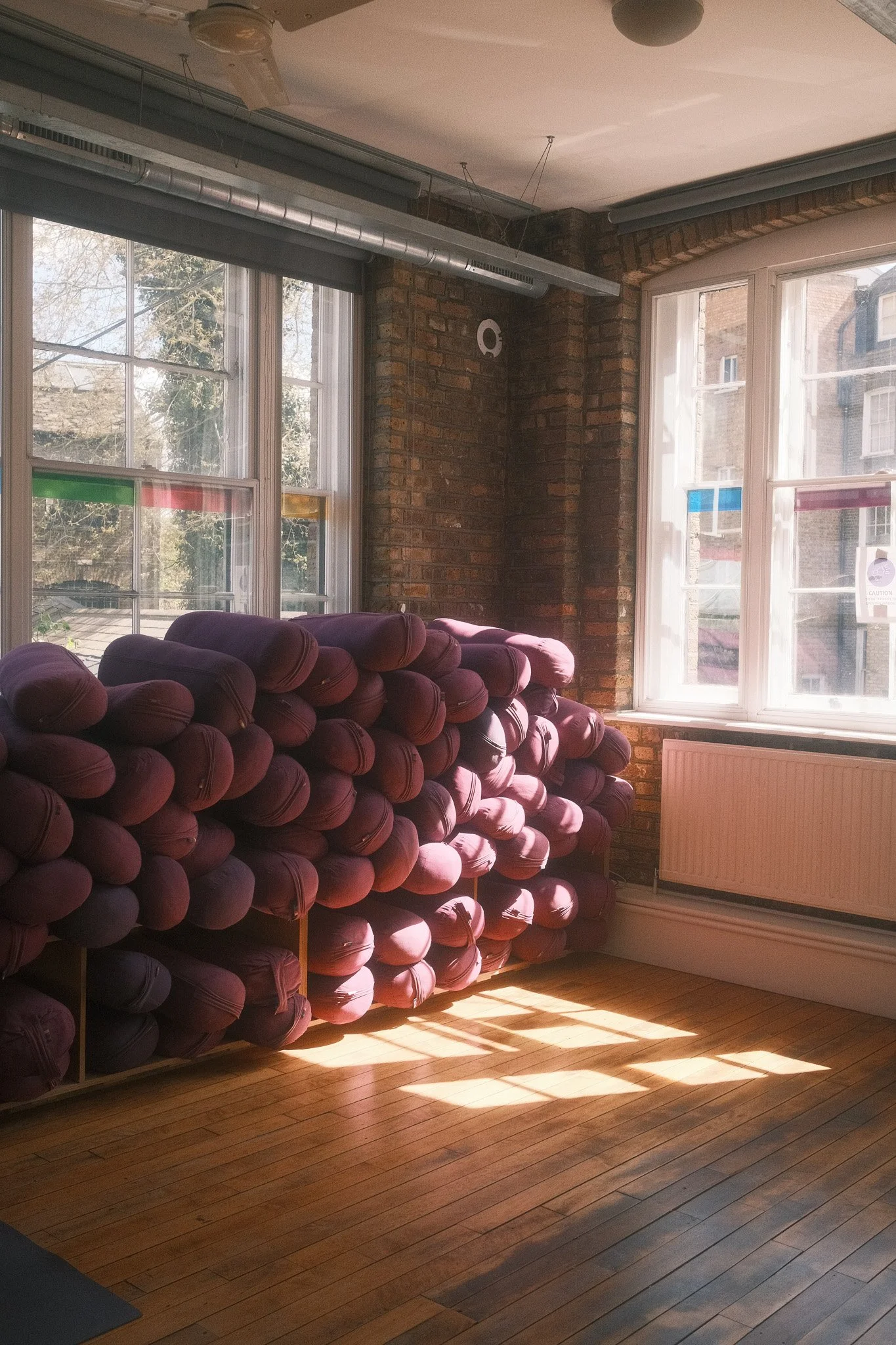 Stacked purple yoga cushions in a sunlit room with brick walls and large windows. triyoga, camden, london