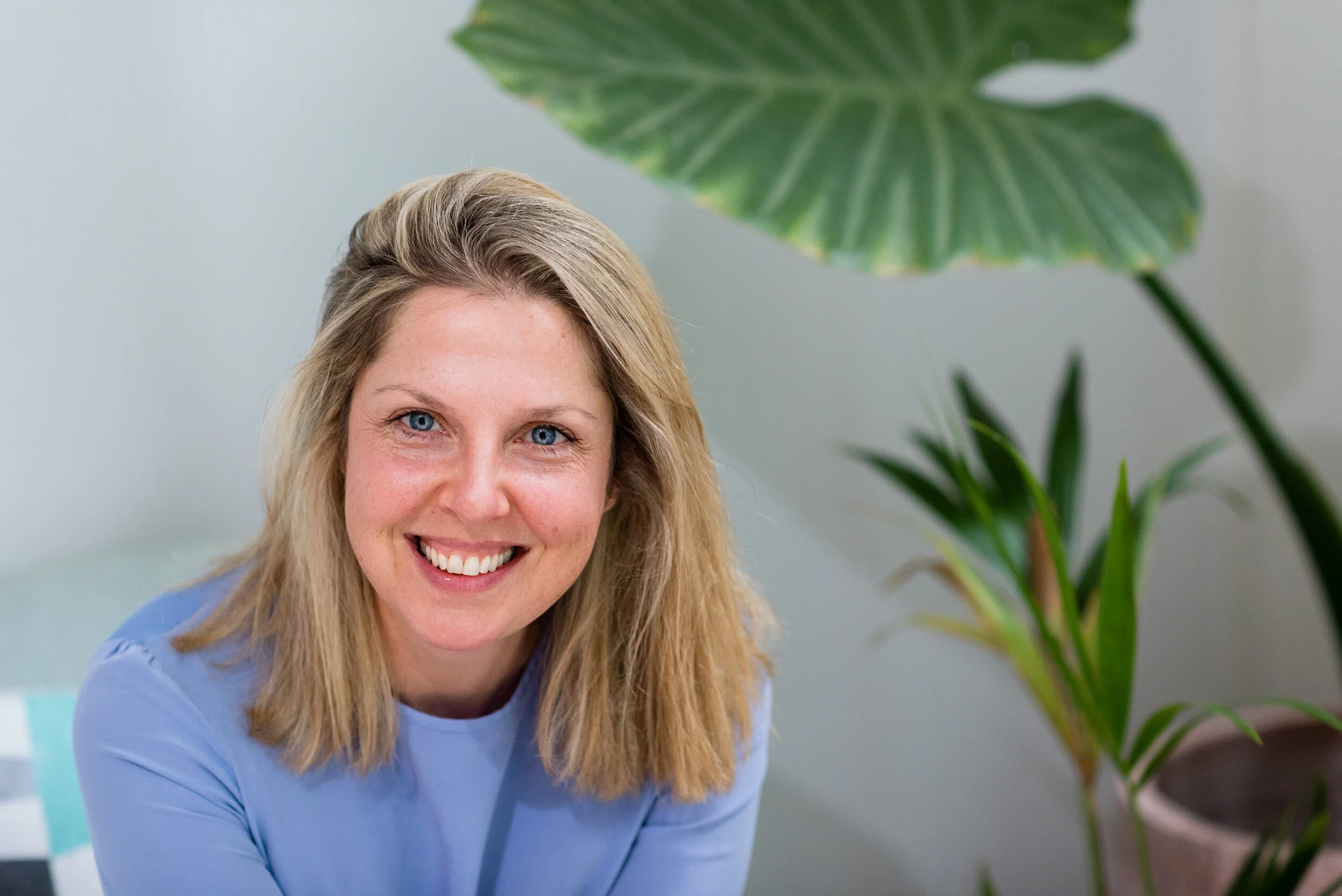 A smiling woman with blonde hair and blue eyes, wearing a light blue top, sitting indoors near a large green plant with broad leaves.