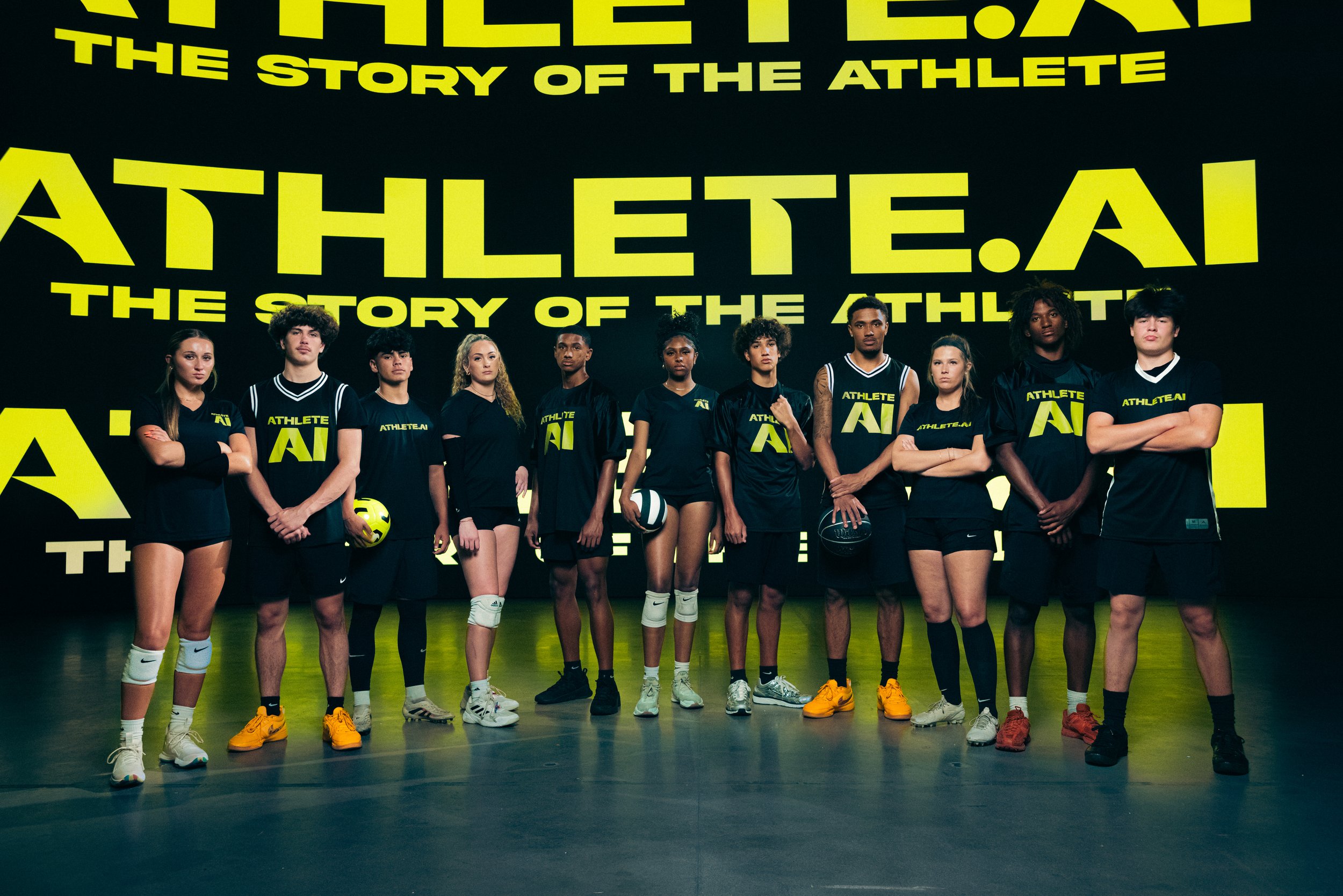 A diverse group of eleven young athletes standing in a line, wearing black athletic clothing with yellow logo and text, against a large black and yellow backdrop with the words 'Athlete.ai' and 'The story of the athlete'.
