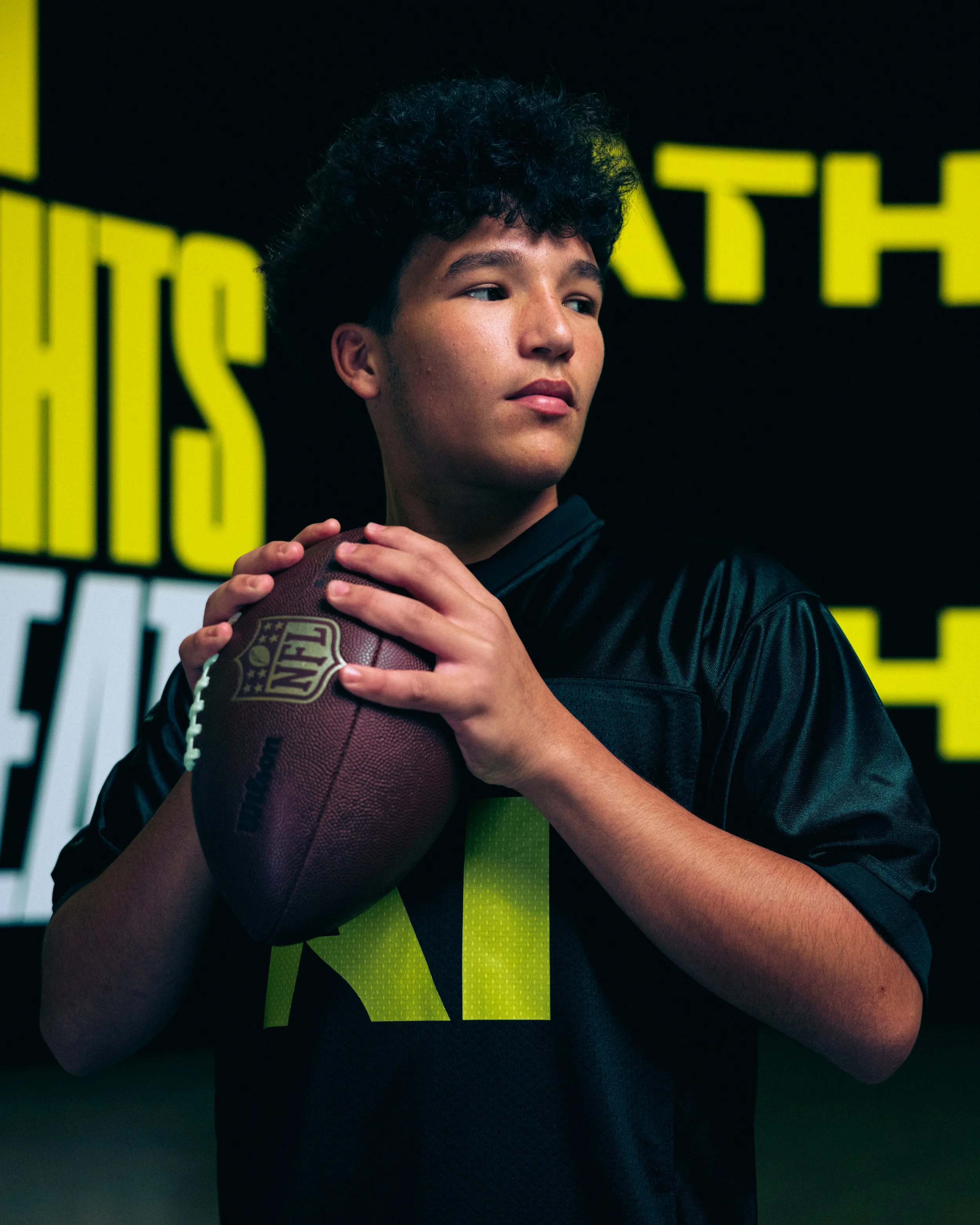 Teenager holding an NFL football, wearing a black sports jersey with a yellow number 11, in front of a yellow and black background.
