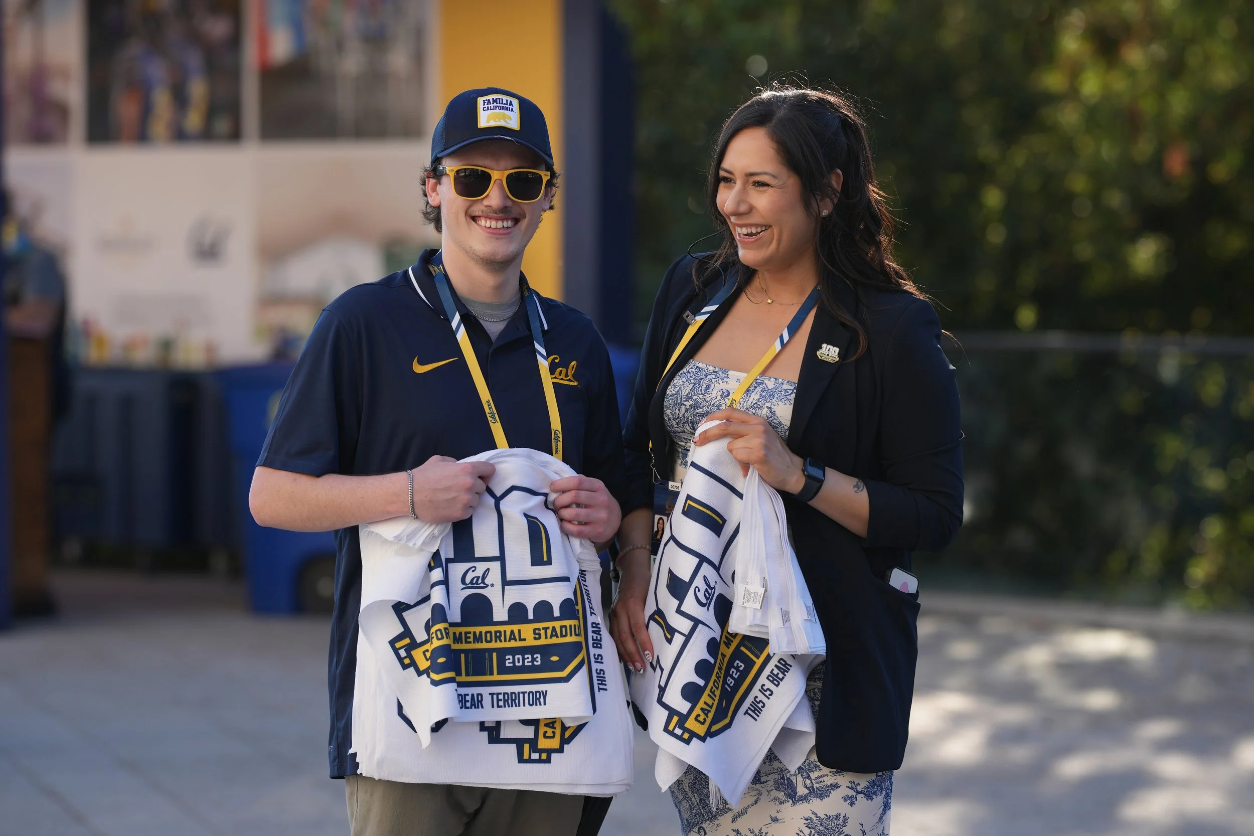 Two people, a young man and a woman, smiling and holding university merchandise, standing outdoors on a campus, with trees in the background.