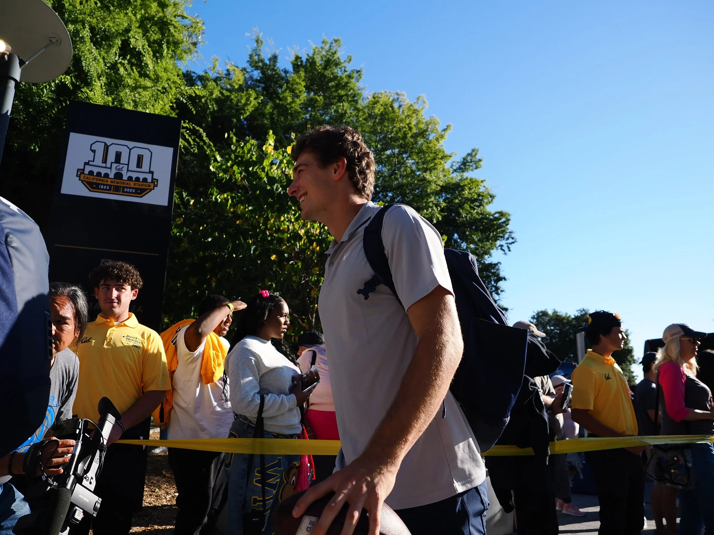 Young man smiling and holding a football, surrounded by people at an outdoor event celebrating the 100th anniversary of California Memorial Stadium, with a sign showing the stadium's logo and the years 1923 and 2023.
