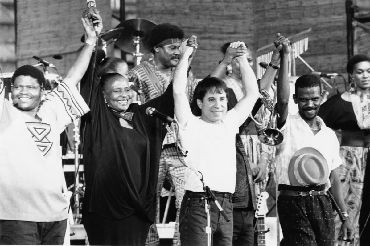 Paul Simon, Miriam Makeba, and members of Ladysmith Black Mambazo pose triumphantly at a concert performance. 