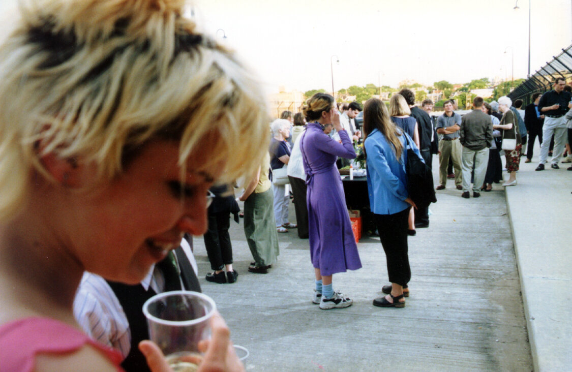 Woman with short blonde hair holding cup in foreground, large crowd of people conversing in groups in background.