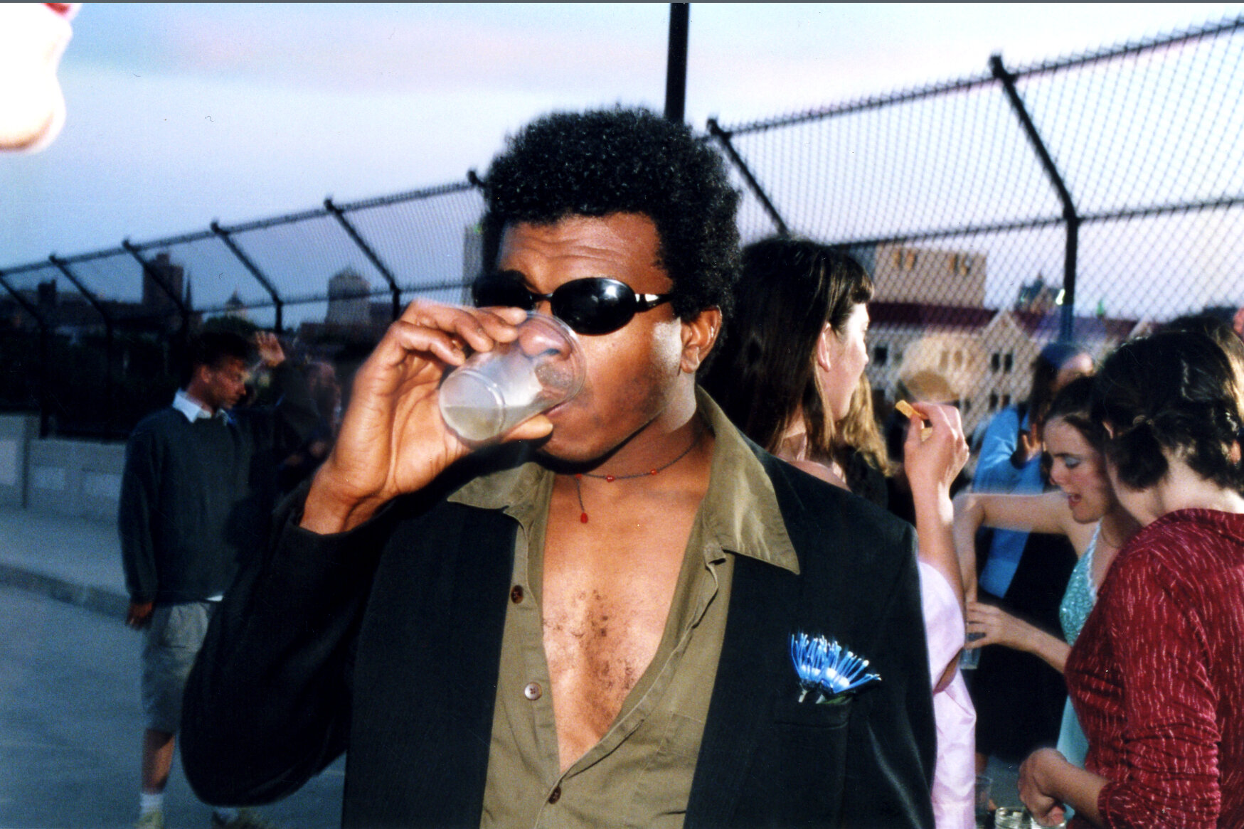 Man in green shirt and black coat with blue flower in pocket and sunglasses drinking from cup, people conversing behind him.