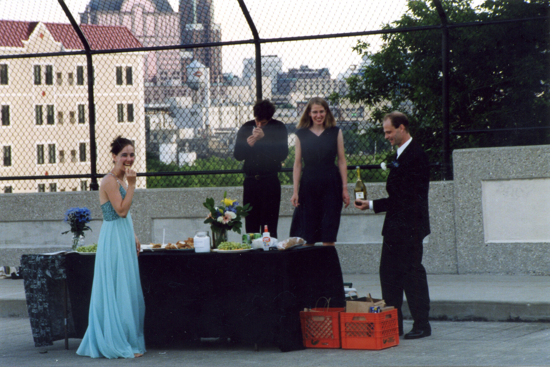 Woman in long blue dress stands next to table, behind it are 2 men and a woman all in black and tall black chain link fence.