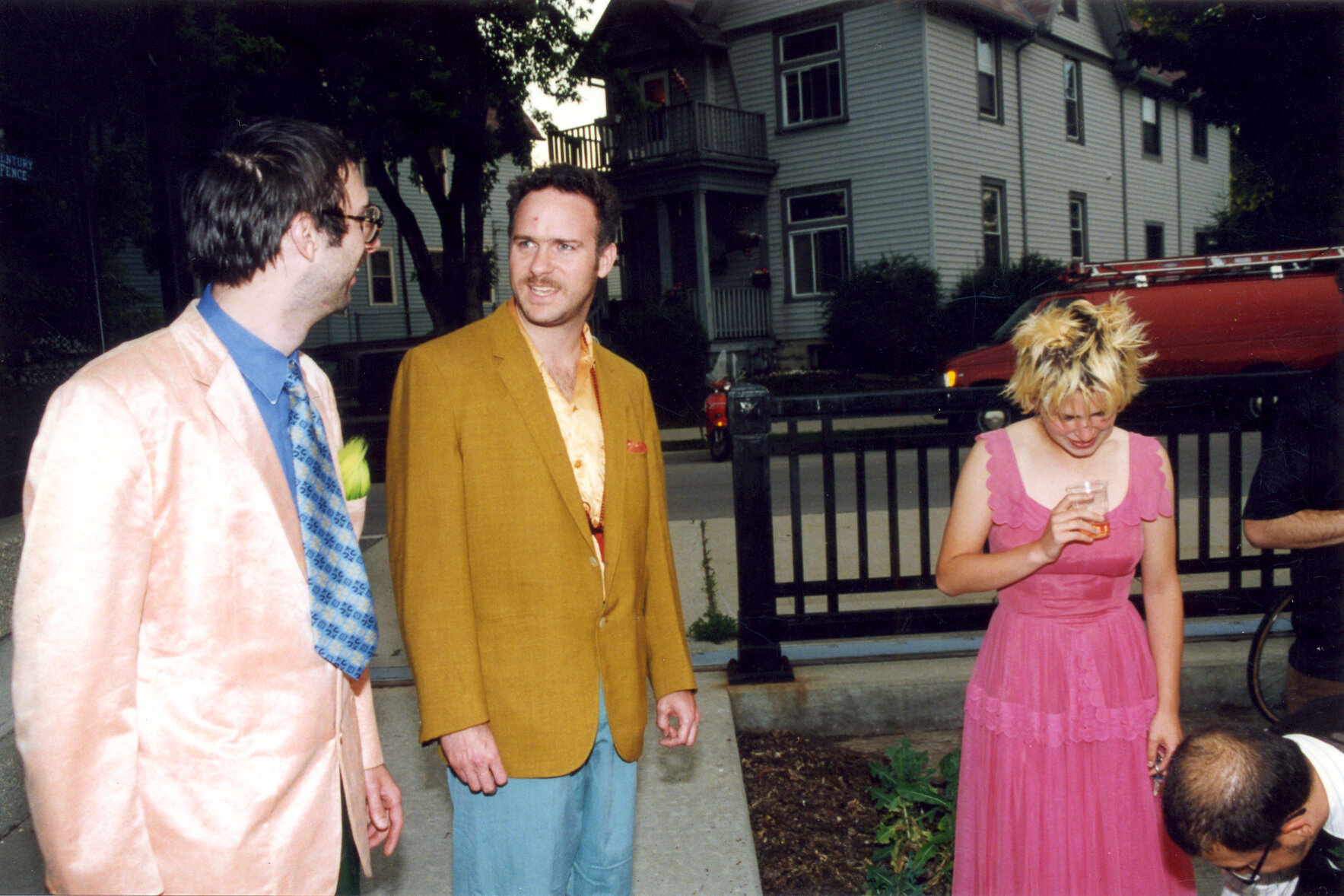 2 men standing next to each other exchange glance, blonde woman in pink dress next to them looks down.