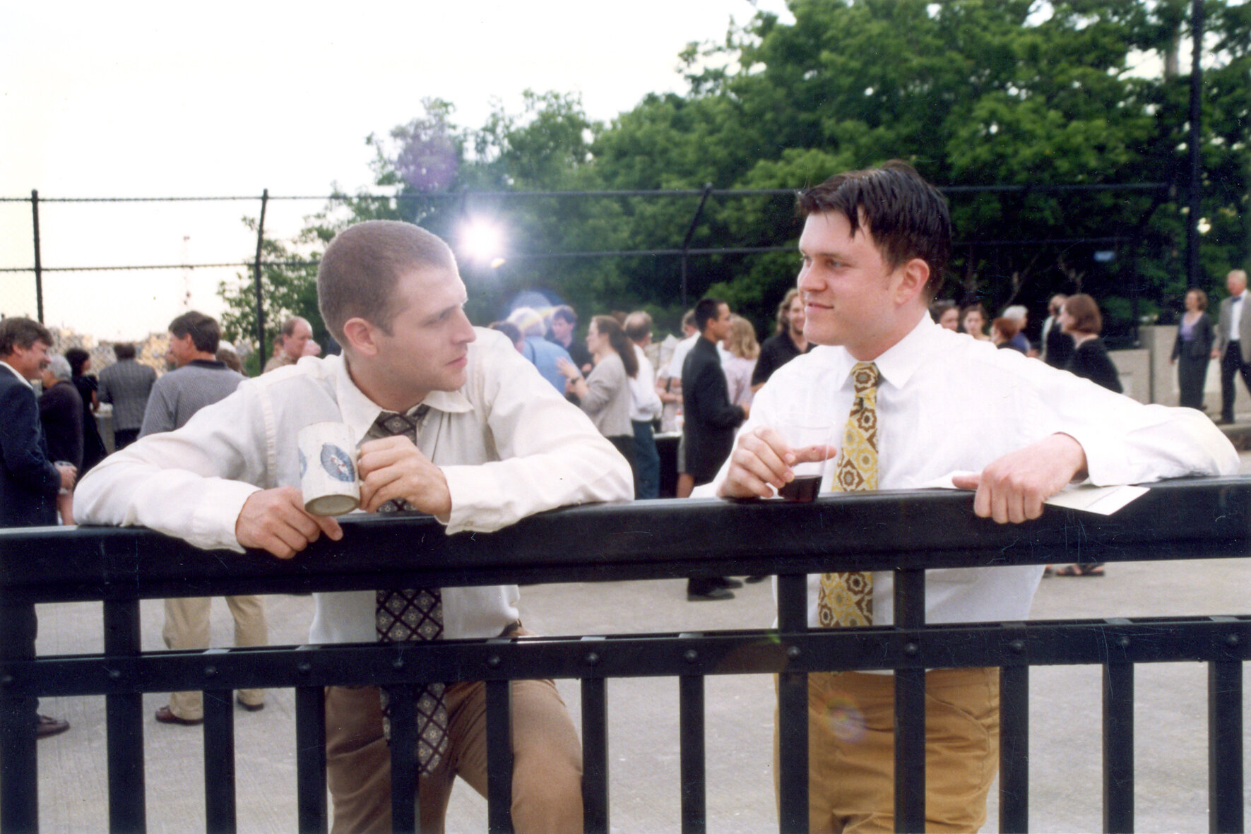 2 men in brown pants and tan shirts lean over black railing while speaking to one another.