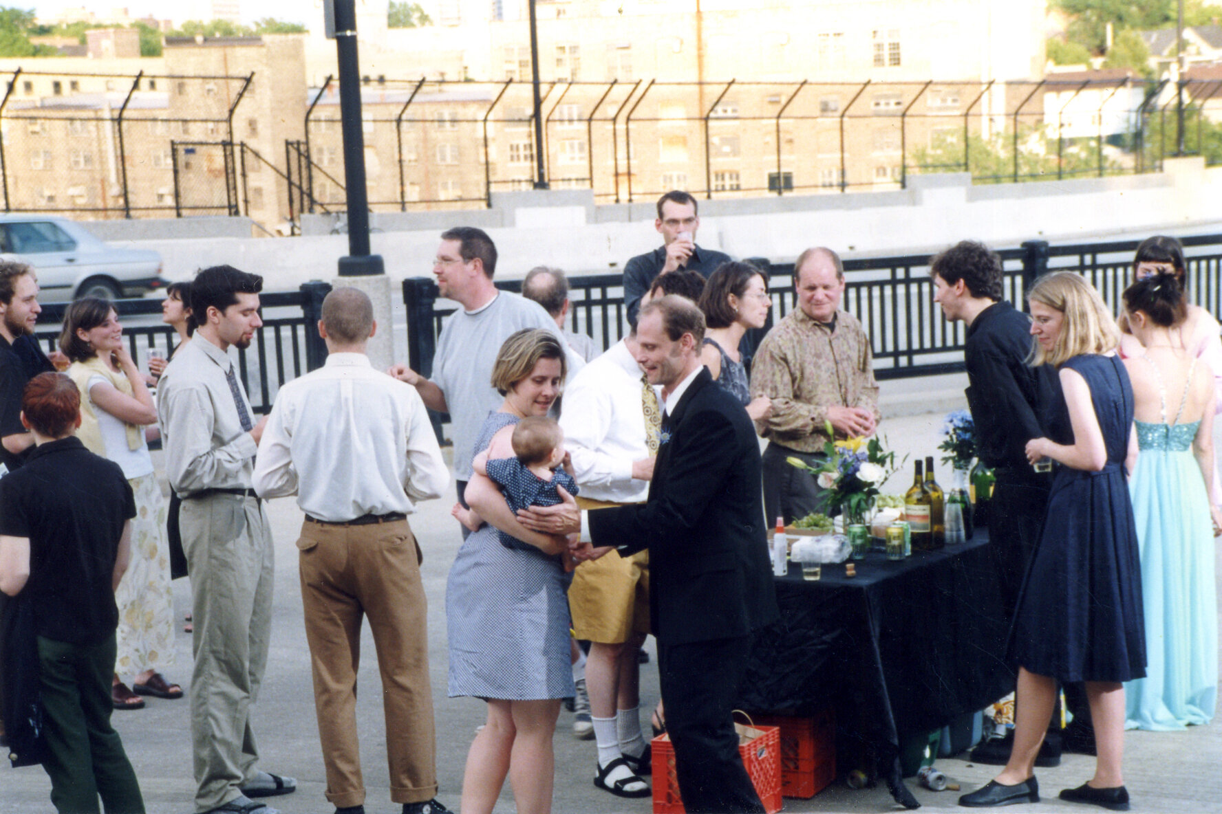 People in formal attire gathered around table with black tablecloth refreshments on it. In foreground, woman hands man baby.