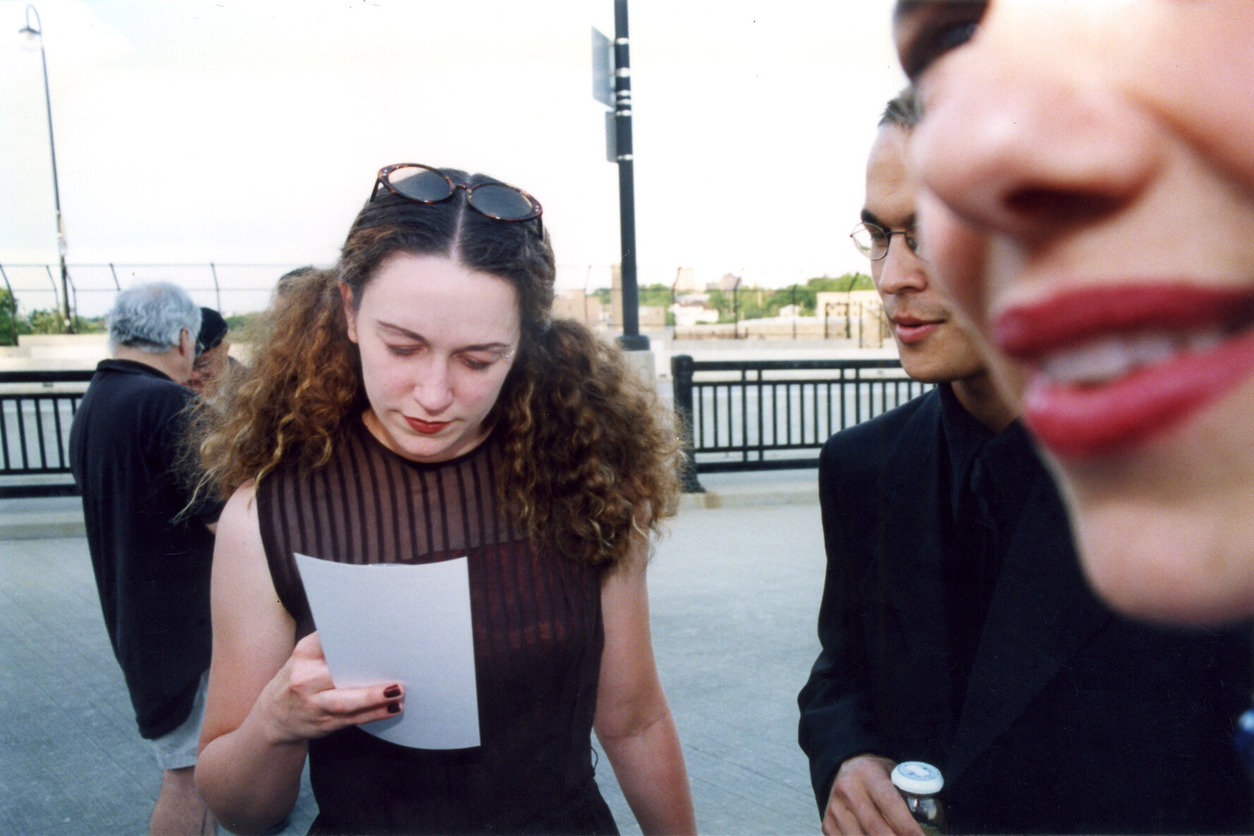 Woman with brown curly hair held back by sunglasses wearing black dress reads pamphlet.