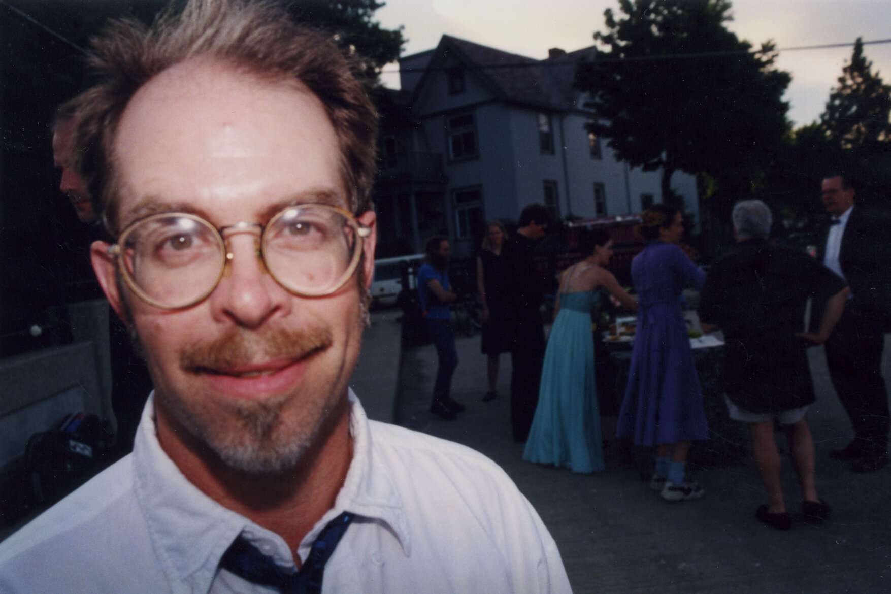 Man in white shirt with dark tie, large round glasses, and brown hair smiles into camera.