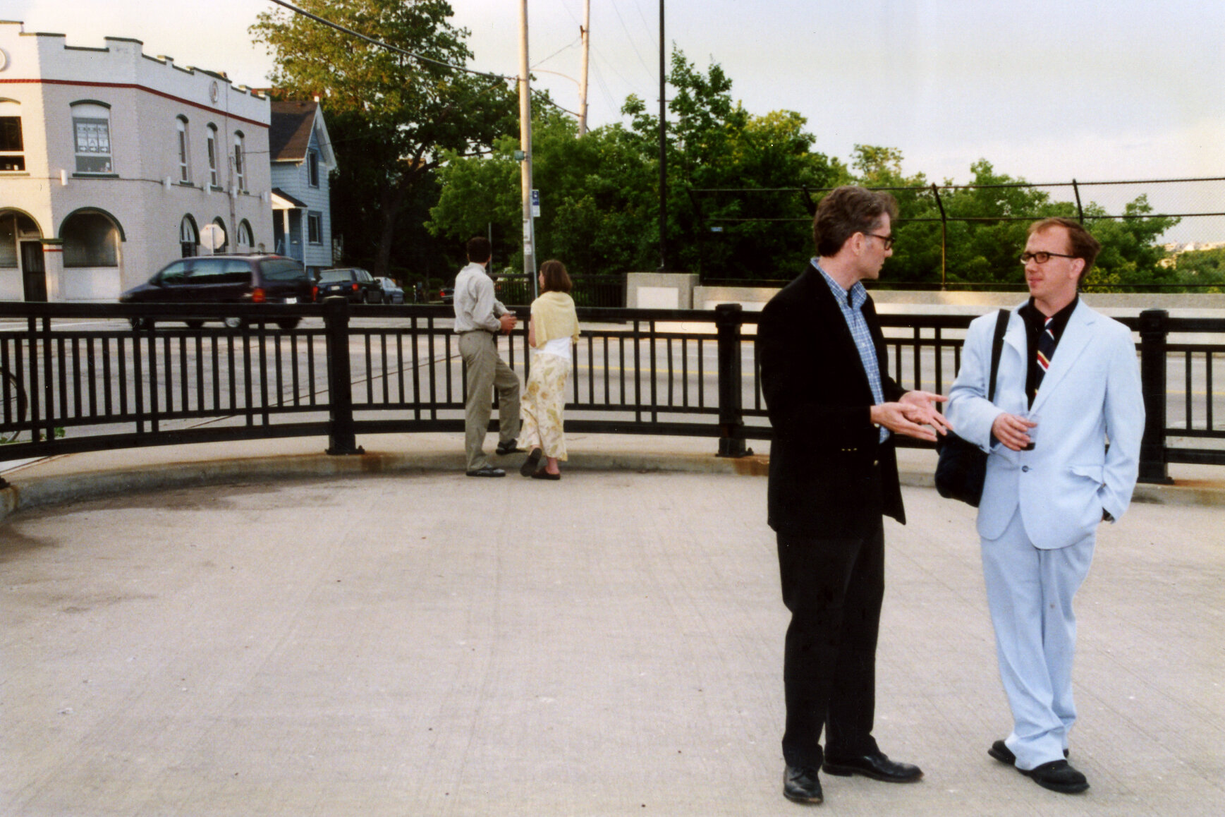 2 men, one in black suit, one in baby blue suit converse in middle of concrete area. 2nd pair stands behind them.